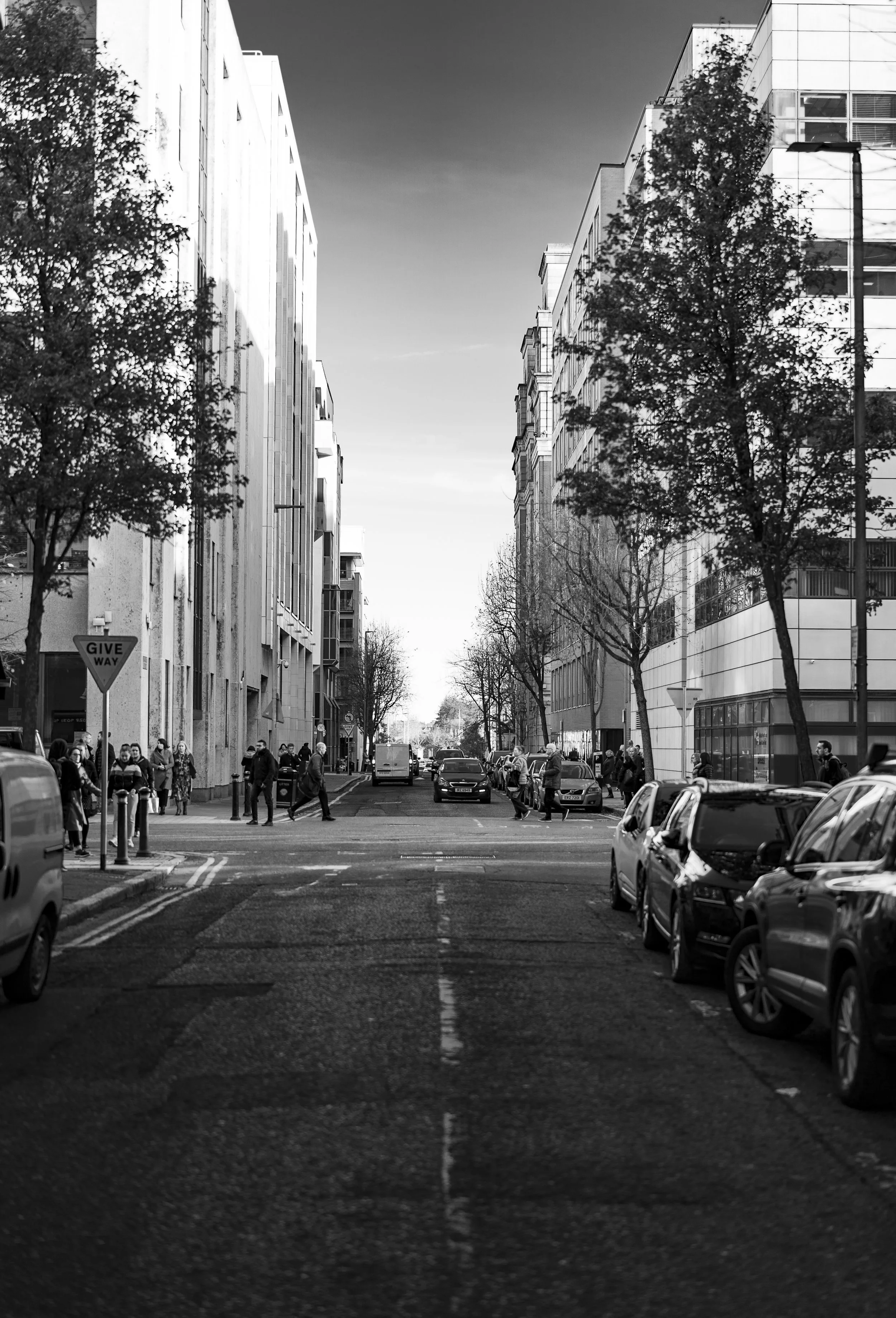 Black and white photo of an urban street lined with tall buildings and parked cars, with pedestrians crossing the street, and a 'Give Way' sign visible.