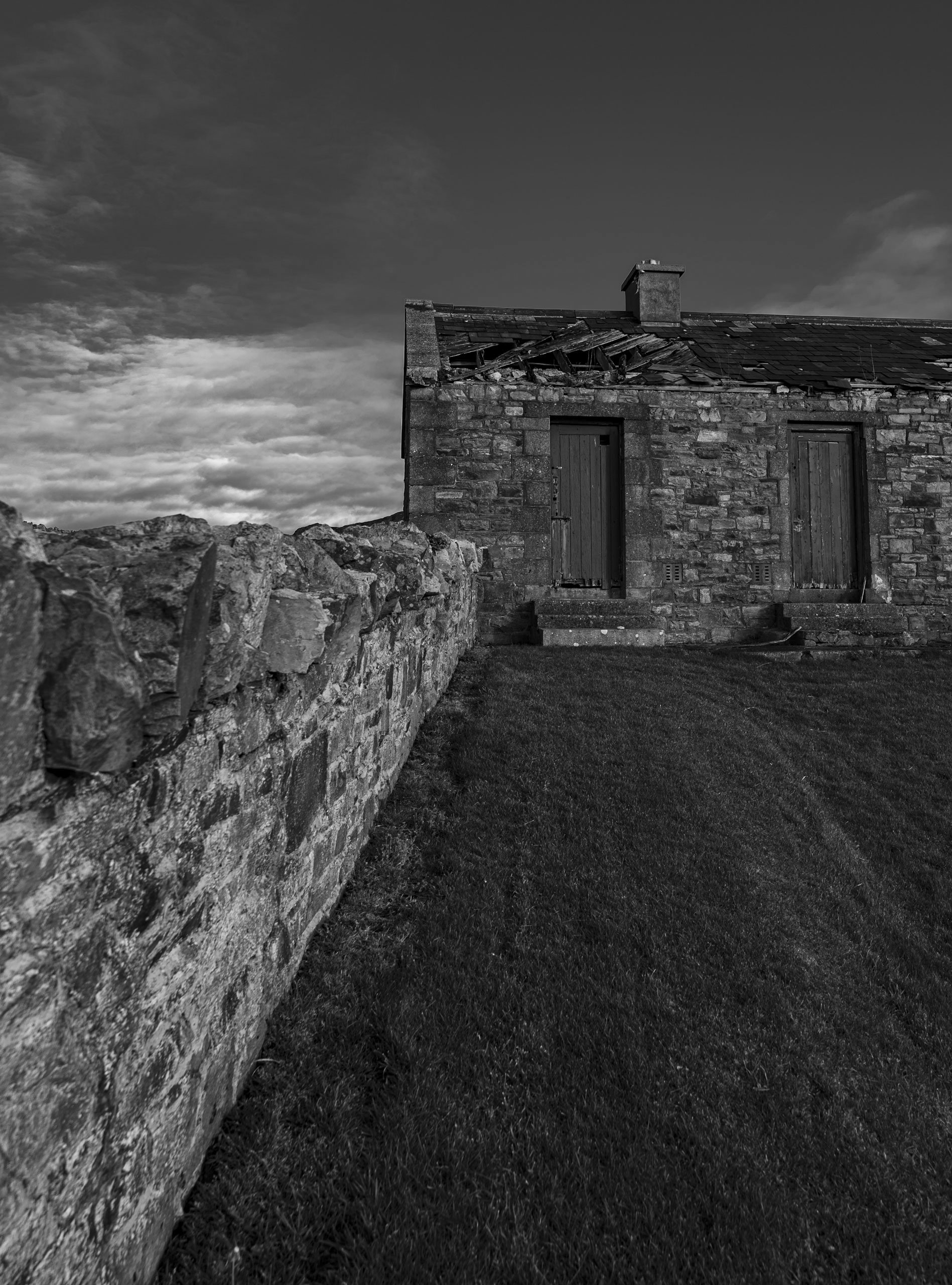 Old stone building with crumbling roof and adjacent stone wall; grassy foreground and cloudy sky background.