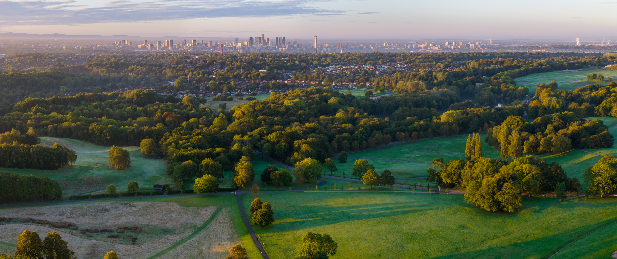 Panoramic aerial image of a sunrise over Heaton Park fields with Manchester skyline and morning light on the horizon.