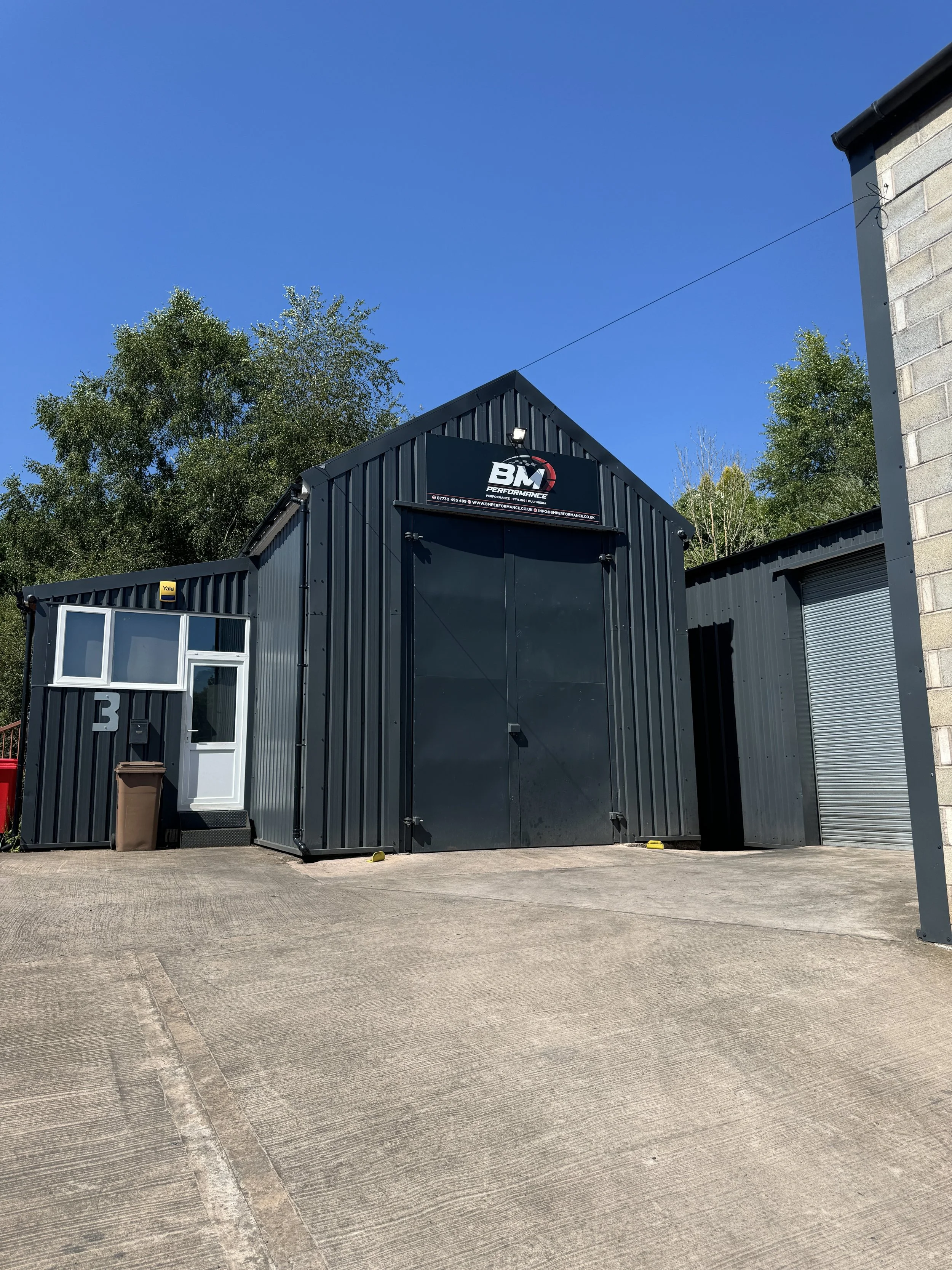 Black industrial building with a large door, a smaller side door, and a window, with a sign for BM Performance on top, and blue sky with trees in the background.