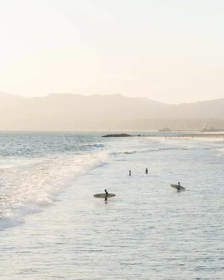 Surfers waiting in the shallow waters of a beach with mountains in the distance during sunset.