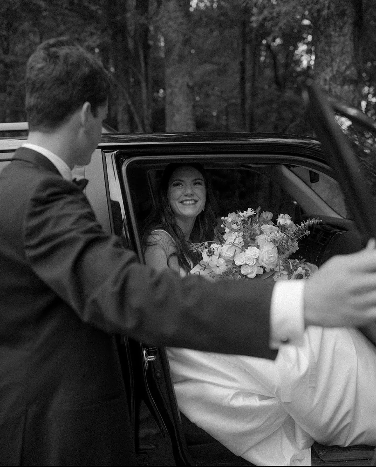 A woman with long hair smiling from the passenger seat of a car holding a bouquet of flowers, while a man in a tuxedo appears to be helping her out of the car in a wooded outdoor setting.