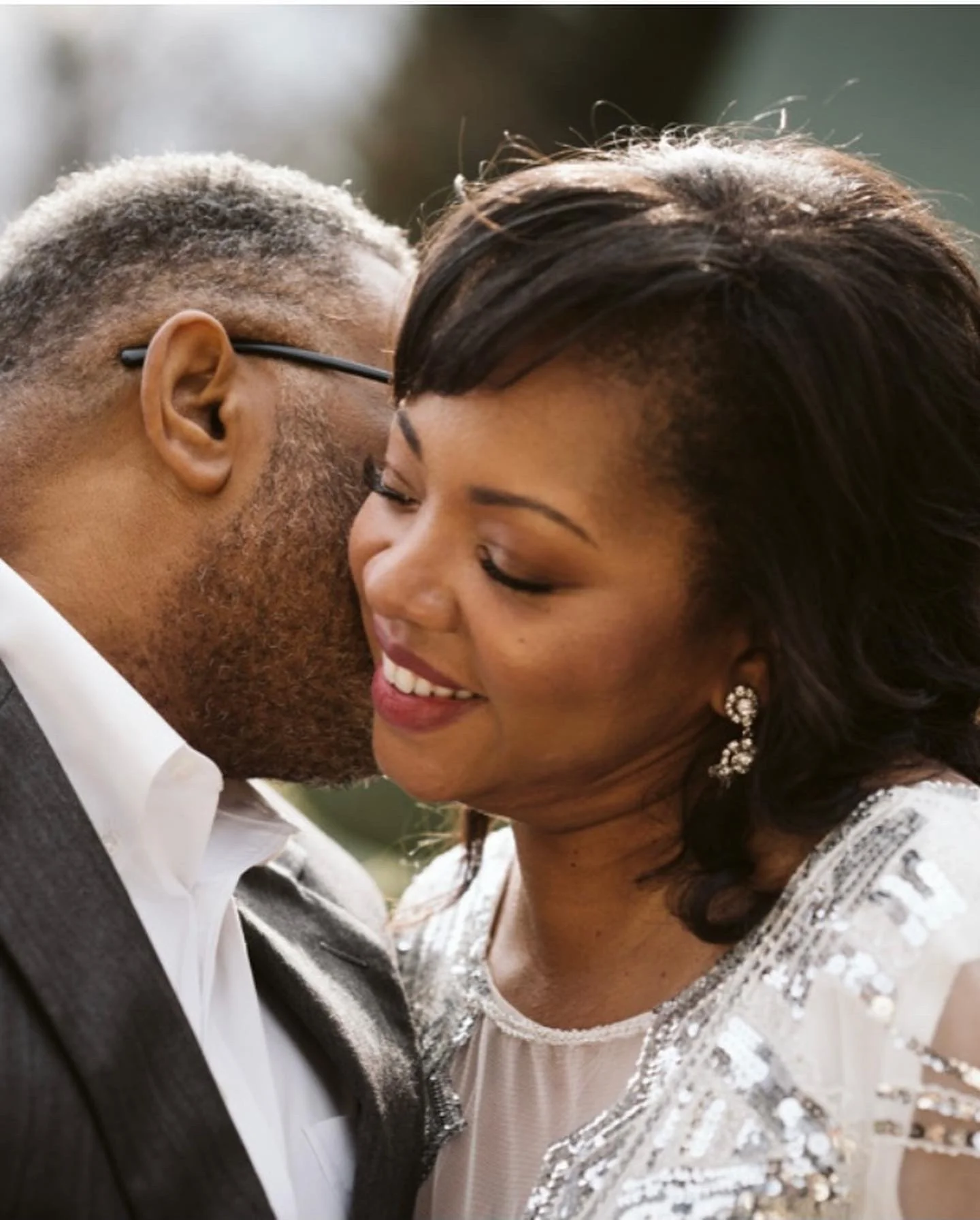 A close-up of a smiling man and woman embracing, with their foreheads touching. The woman has dark hair and earrings; the man has gray hair and wears glasses. They are dressed formally, suggesting a special occasion.