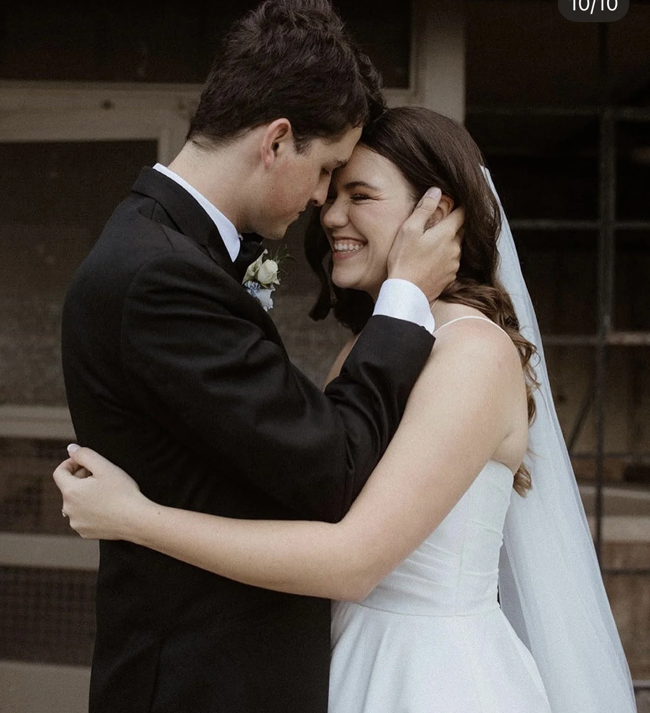 A bride and groom embrace, smiling, with the groom in a black tuxedo and the bride in a white wedding dress, indoors.