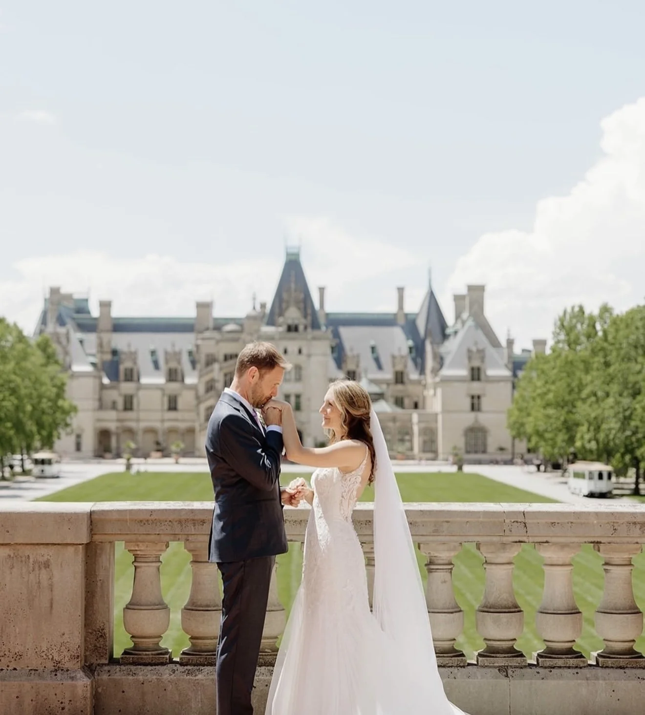 A bride and groom stand on a balcony in front of a castle, holding hands and smiling at each other on their wedding day.