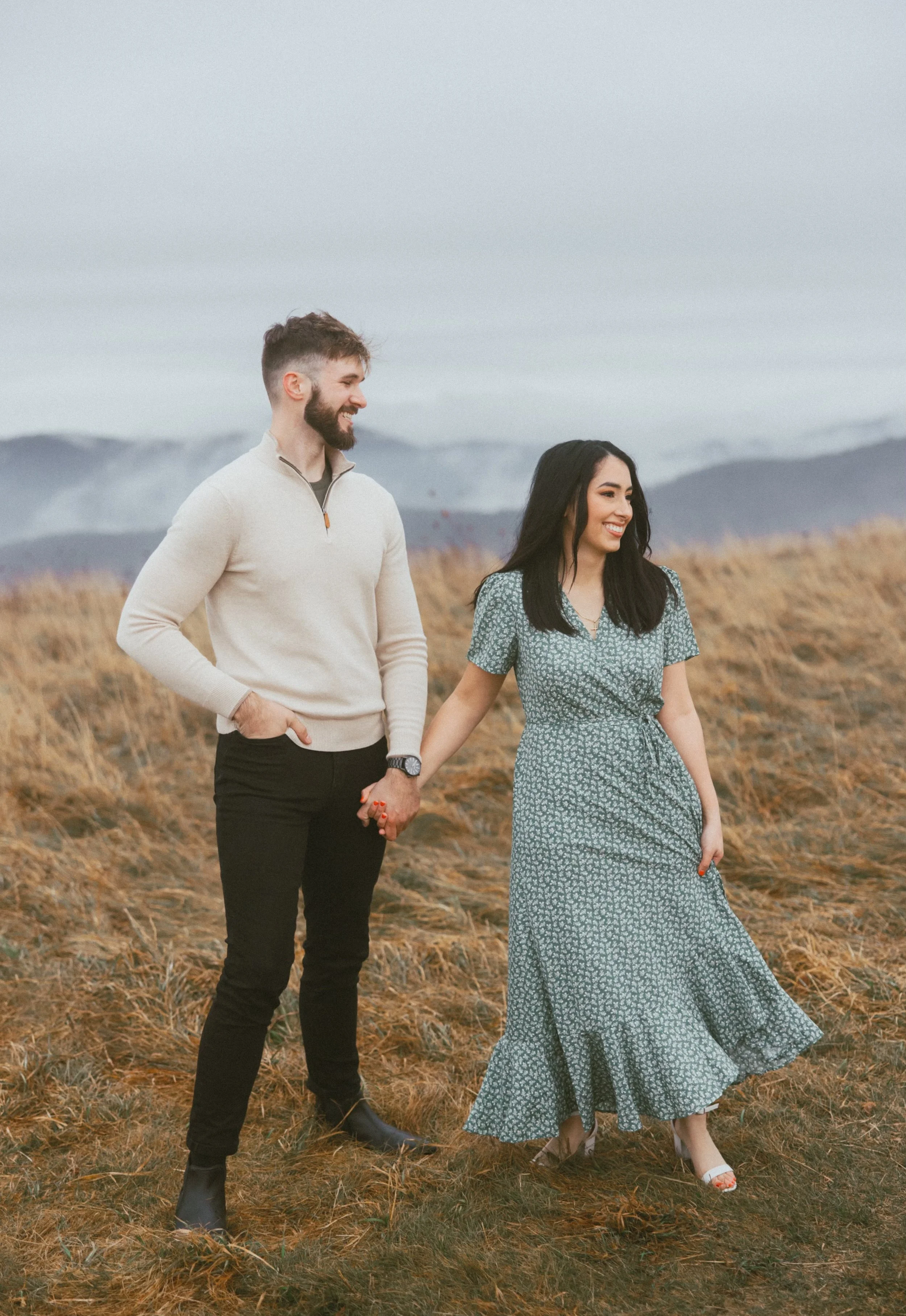 A man and woman holding hands and walking through a field with mountains in the background, smiling and looking at each other.