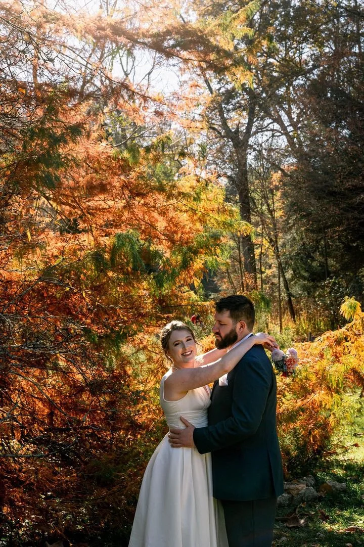 A bride and groom hugging outdoors among trees with autumn foliage.