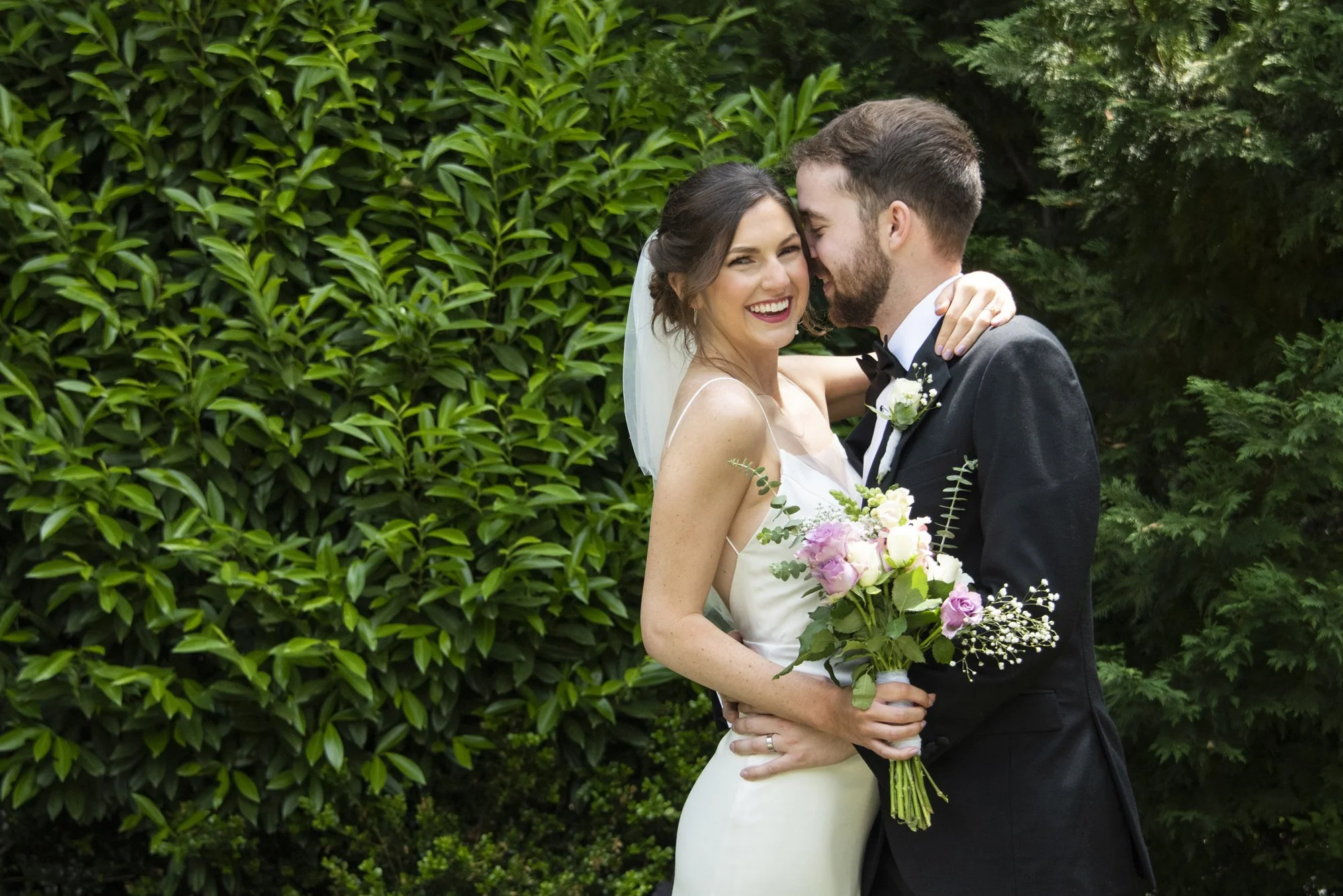 A bride and groom sharing a kiss outdoors, with the bride holding a bouquet of pink and white flowers, surrounded by green foliage.