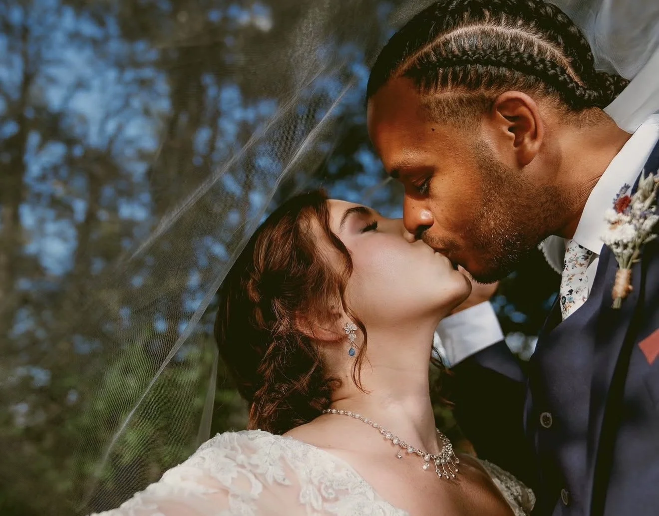 A bride and groom sharing a kiss under a veil outdoors during their wedding.