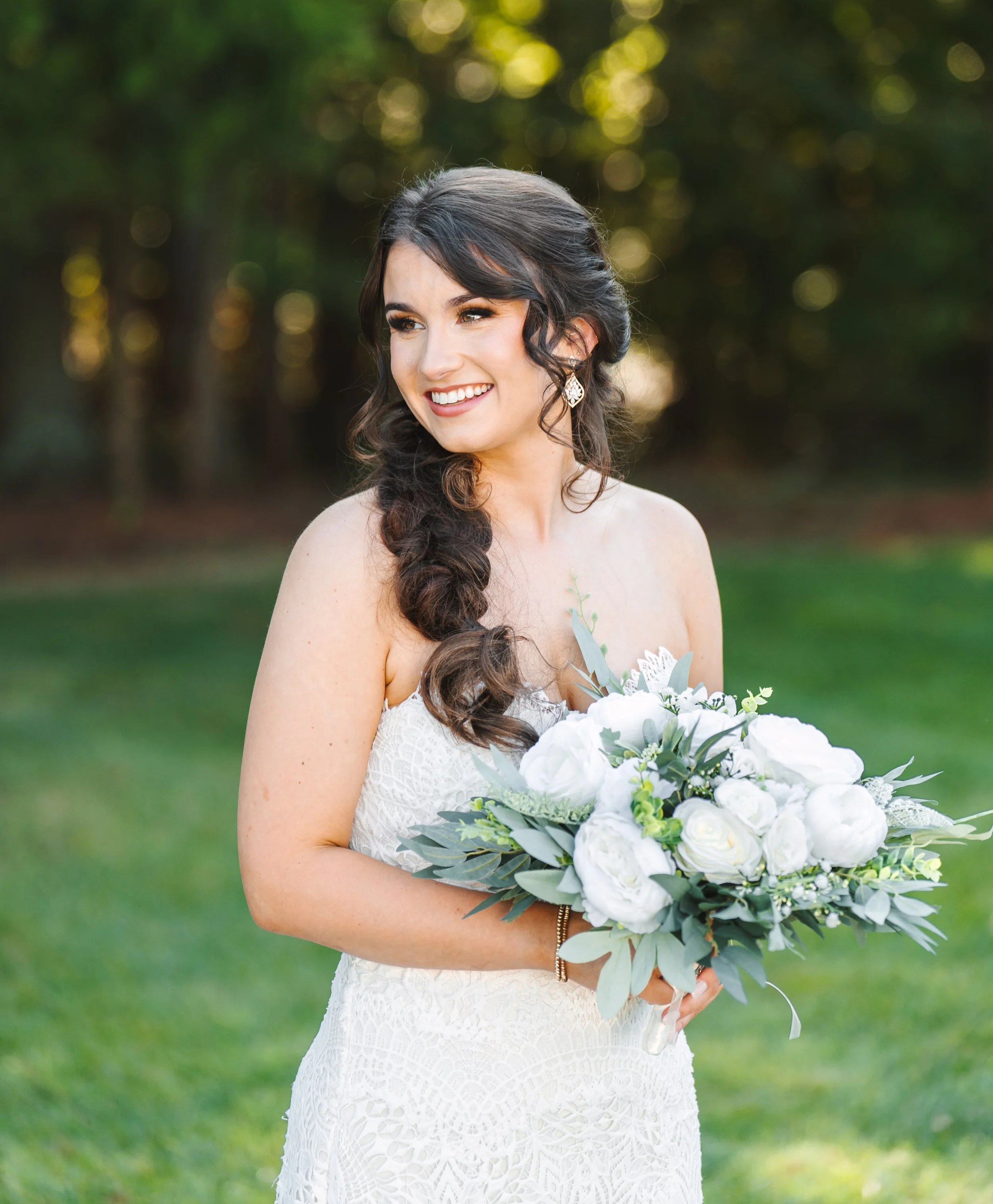 A smiling woman with dark hair in loose curls, wearing a white lace dress and ornate drop earrings, holding a bouquet of white flowers and greenery, outdoors with green trees and bokeh sunlight in the background.