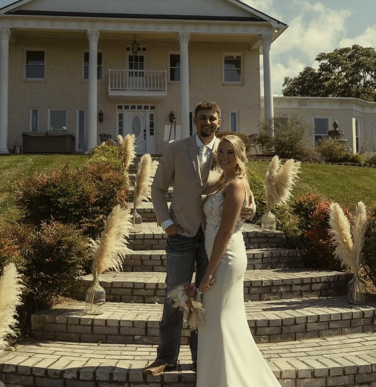 A smiling couple in wedding attire posing on a brick staircase outside a large, historic house. The man is wearing a light-colored suit, and the woman is in a white wedding gown holding a bouquet.