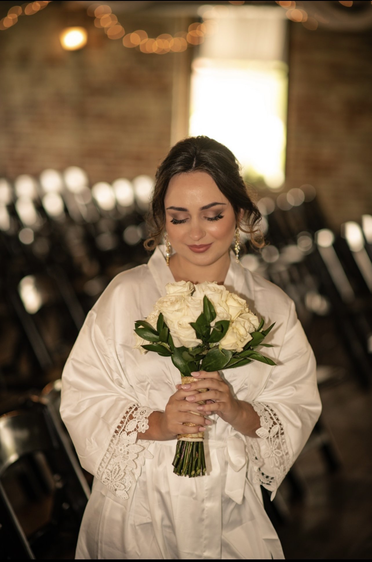 A woman with dark hair and earrings, wearing a white lace-trimmed robe, holding a bouquet of white roses, standing in a room with blurred chairs and string lights in the background.