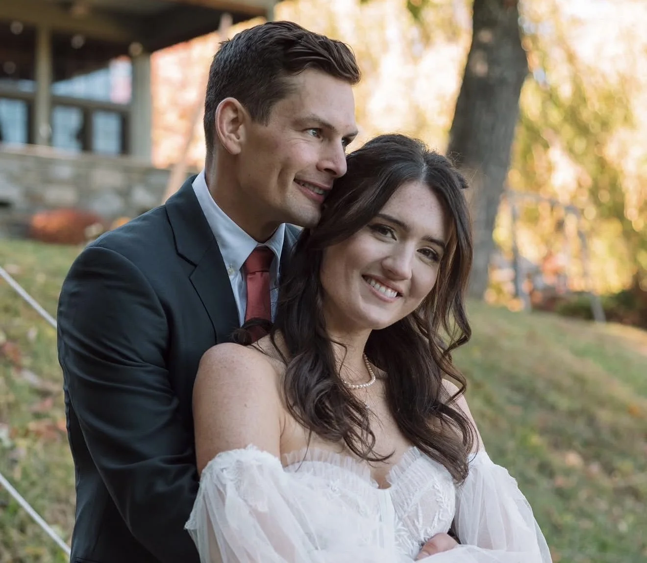 A smiling couple, a man in a dark suit and a woman in a white dress, standing outdoors in a park with fall foliage.