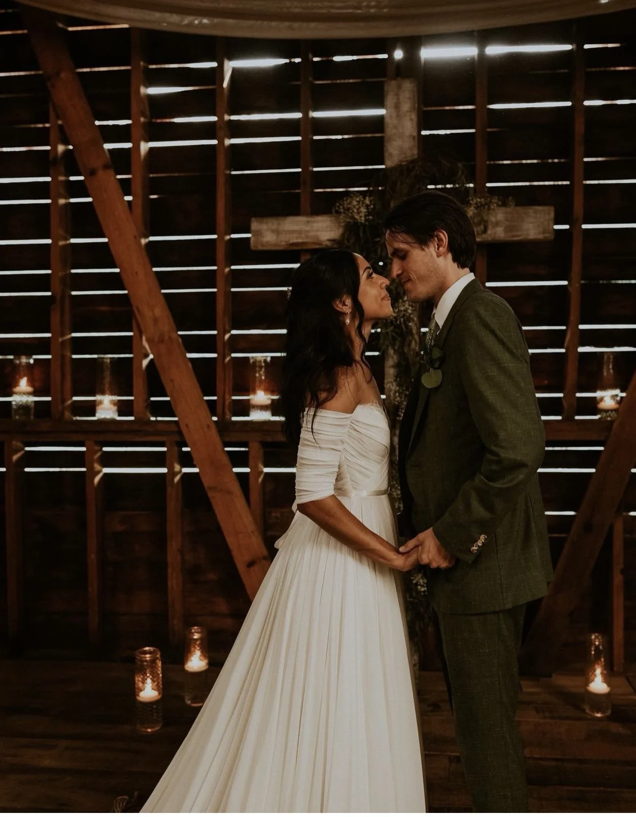 A bride and groom holding hands, standing close with foreheads touching inside a rustic wedding venue with wooden walls and candles.