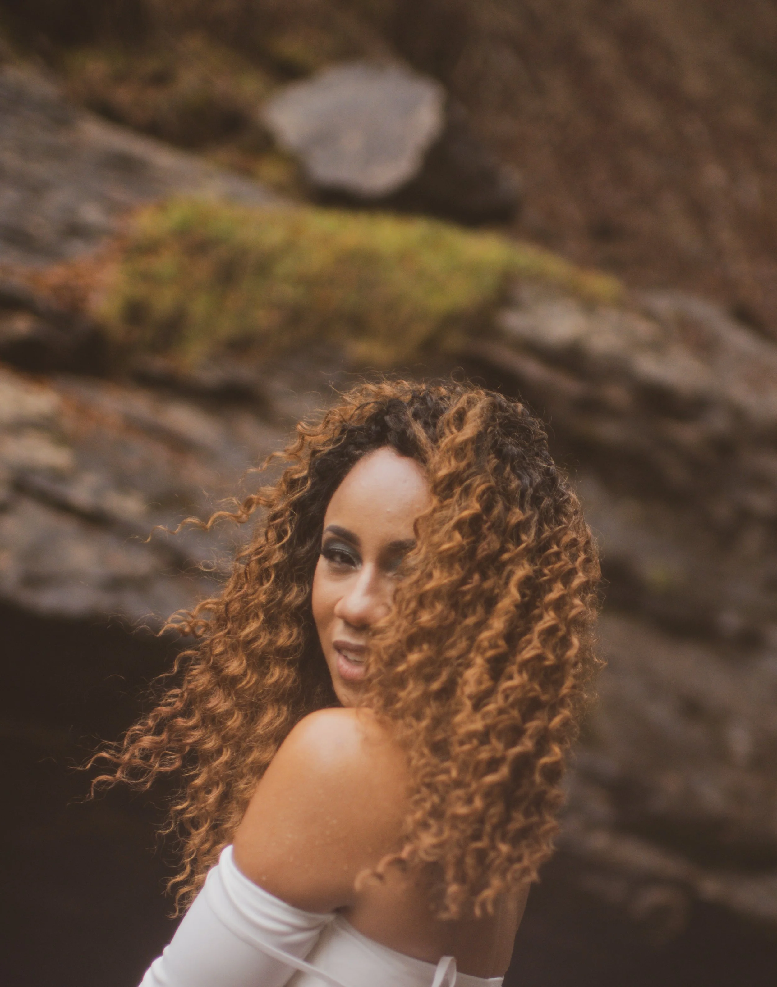 A young woman with dark, curly hair posing outdoors against a colorful, graffiti-covered wall with green vines.