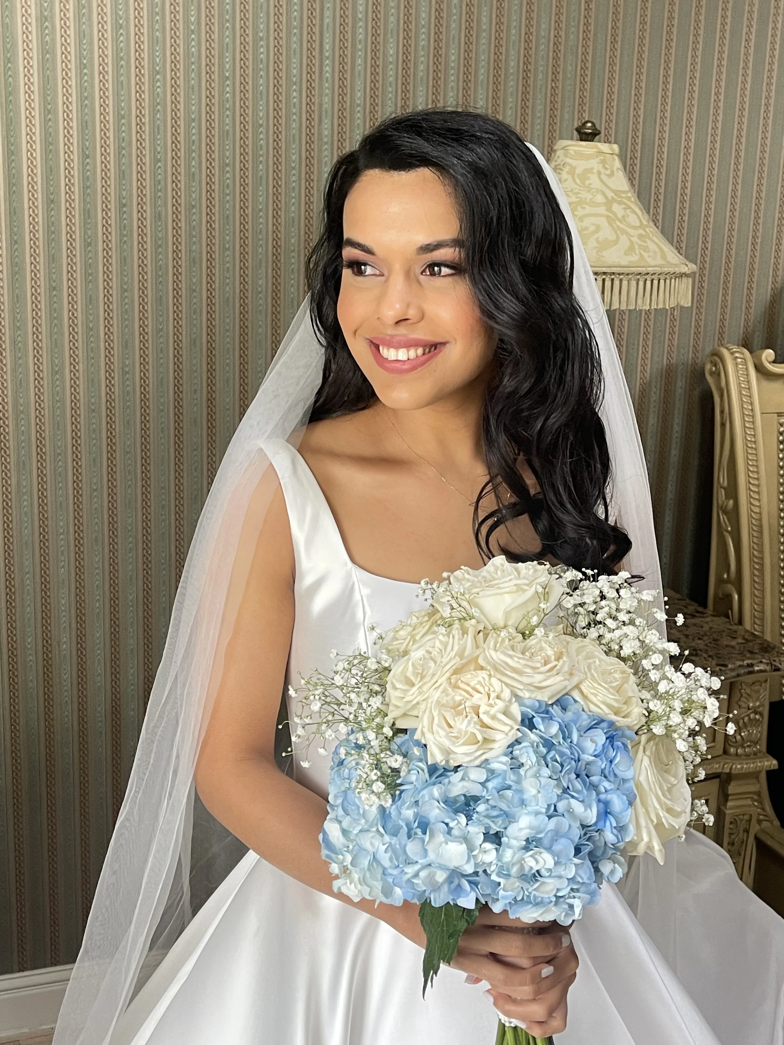 A bride with dark curly hair and makeup, wearing a white wedding dress and veil, holding a large bouquet of blue and white flowers.