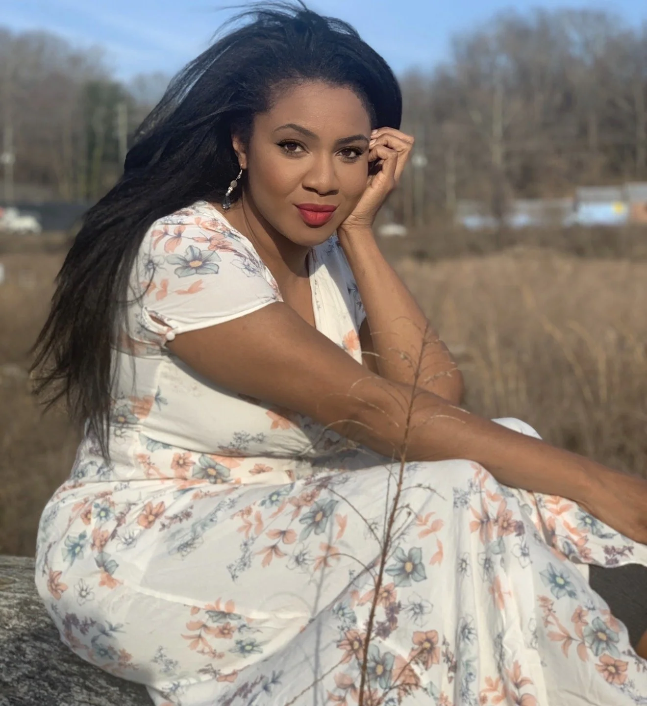 A woman with long black hair sitting outdoors on a rock, wearing a white floral dress. She is resting her head on her hand and looking at the camera, with a field and trees in the background.