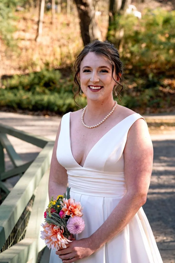 A young woman in a white wedding dress is smiling and holding a colorful bouquet of flowers. She is standing outdoors on a bridge with trees and foliage in the background.