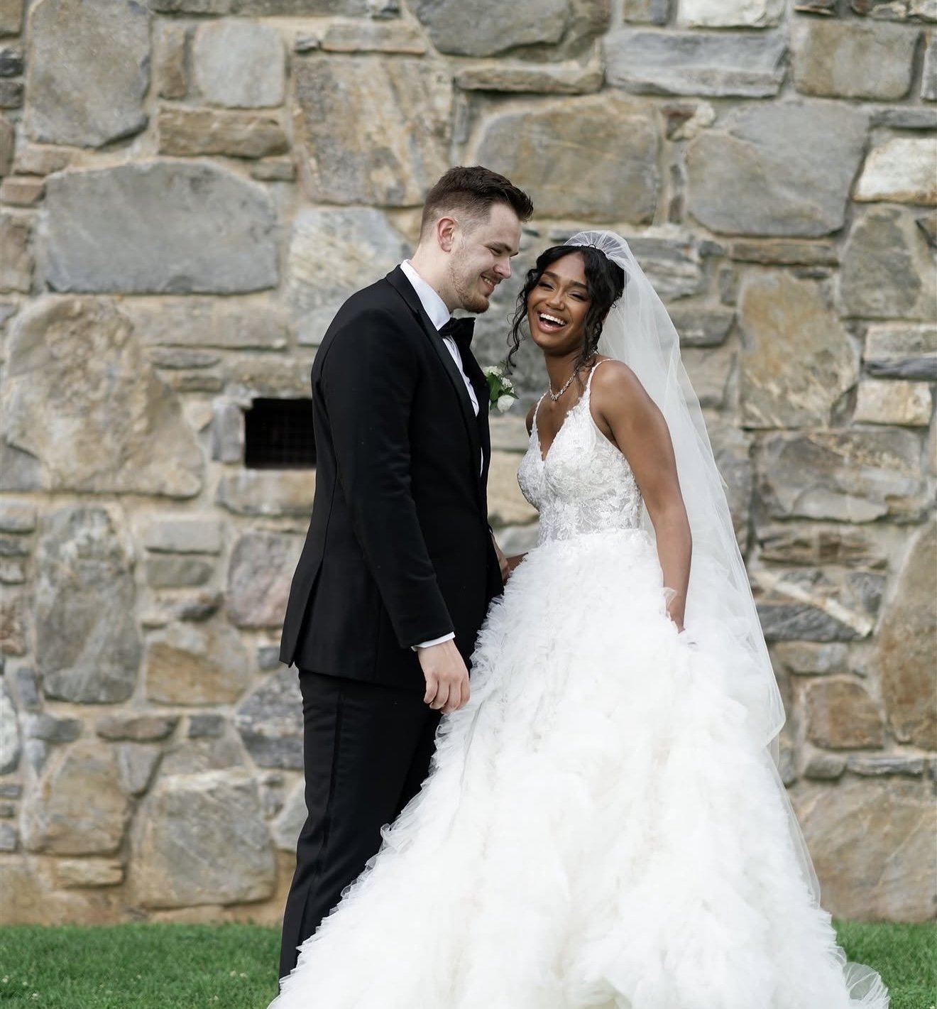 A happy bride and groom are standing close together outdoors against a stone wall, smiling and laughing, on their wedding day. The bride wears a white wedding gown with lace details and a veil, while the groom is dressed in a black tuxedo with a bow 