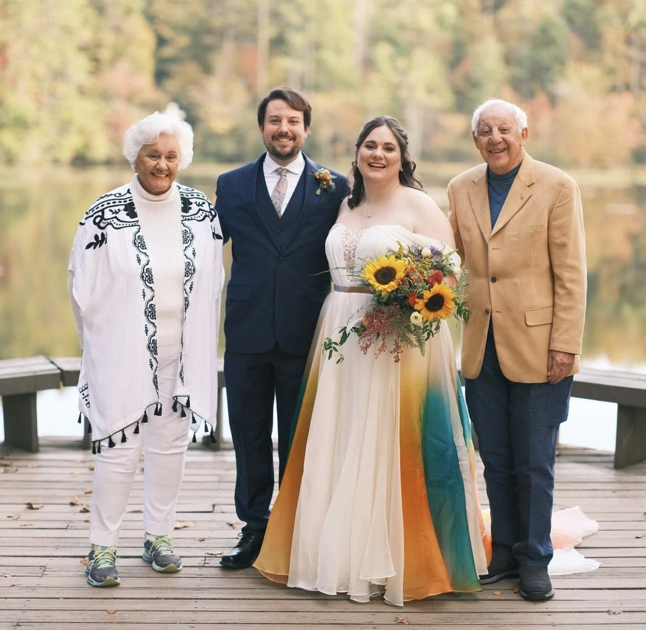 A wedding party outdoors by a lake with trees in fall colors. The bride in a floral rainbow-colored gown holds a sunflower bouquet, standing between a groom in a navy suit and two older adults, one in a cream and black embroidered jacket and white pa