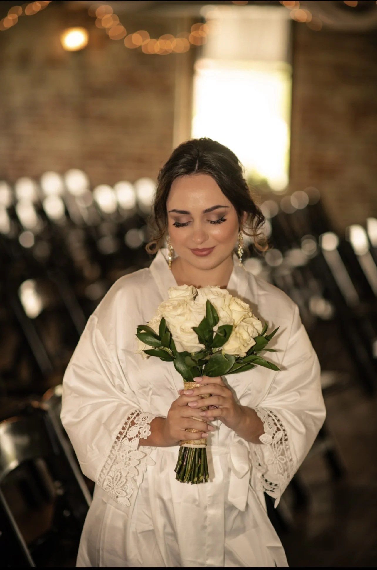 A smiling woman in a white lace wedding dress holding a bouquet of daisies, standing outdoors with lush green foliage in the background.