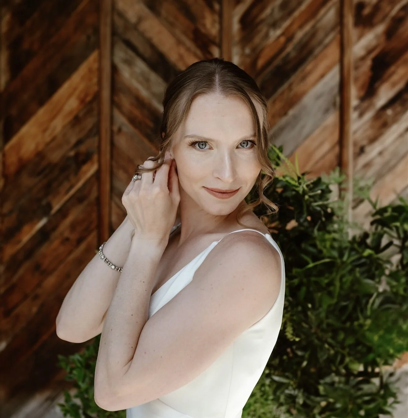 A woman with fair skin and blue eyes wearing a white dress, smiling gently while touching her ear, standing in front of a wooden and green foliage background.