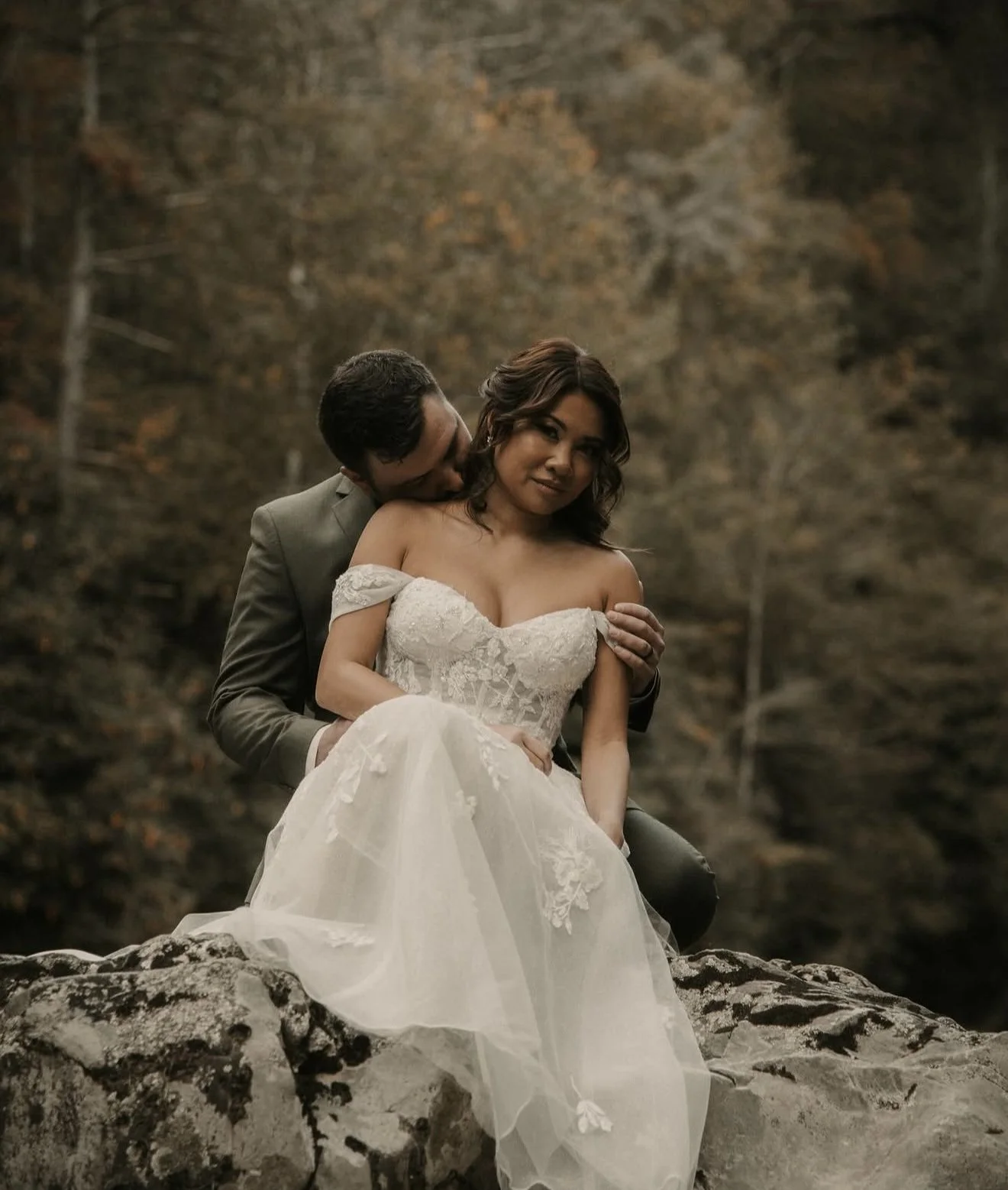 A man and woman in wedding attire sitting on a rock outdoors, with the man kissing the woman on the cheek. The woman is wearing a white off-the-shoulder wedding dress, and the man is in a gray suit. They are surrounded by autumn trees in the backgrou