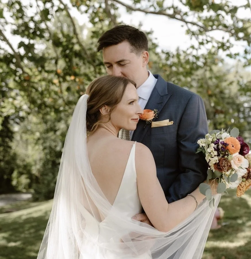 A bride and groom embrace outdoors during their wedding. The bride holds a bouquet of colorful flowers and wears a white sleeveless dress with a veil. The groom wears a navy suit with a boutonnière.