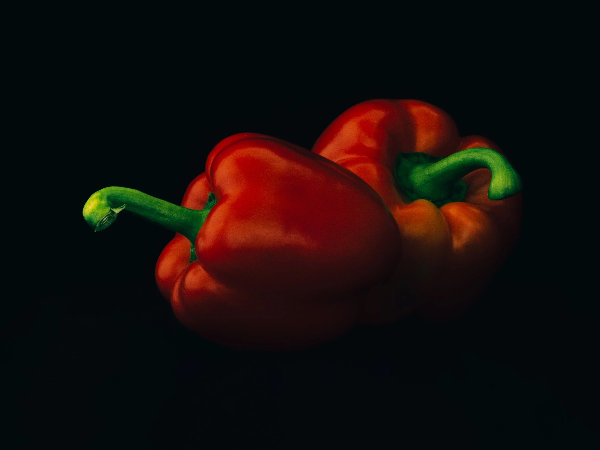 Twee rode paprika's met groene stelen, liggend op een zwarte achtergrond.