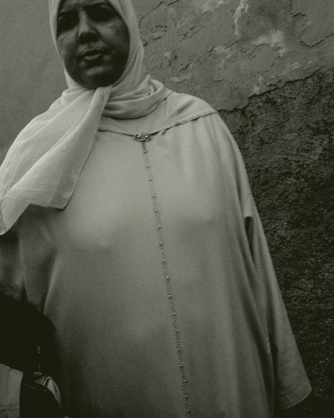 Een vrouw gekleed in een lichte top en een hoofddoek, staand voor een ruwe muur, op een zwarte en witte foto.