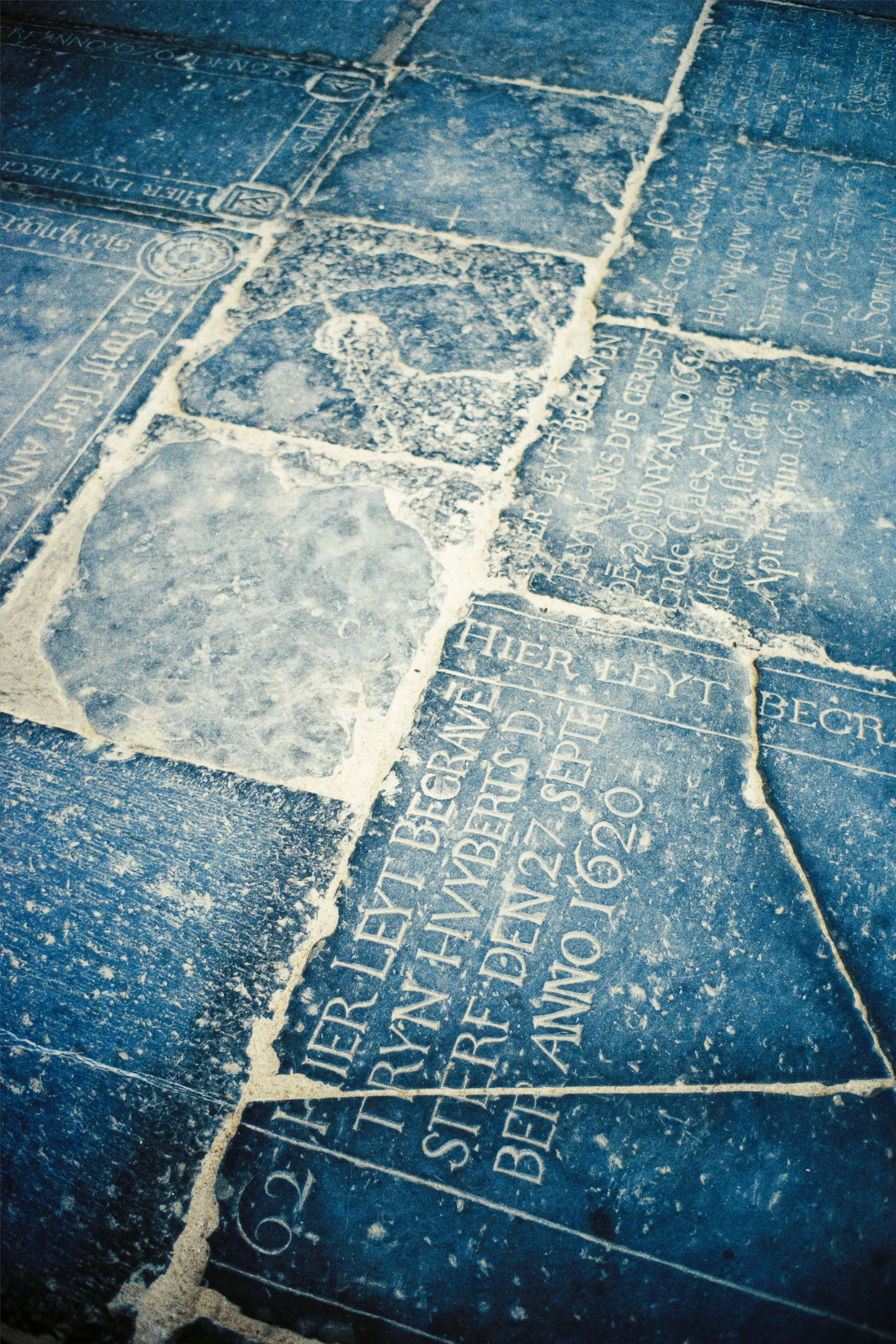 Ingewassen grafstenen met inscripties op de vloer, sommige hard en andere zacht, waarschijnlijk in een kerkhof of historisch monument.