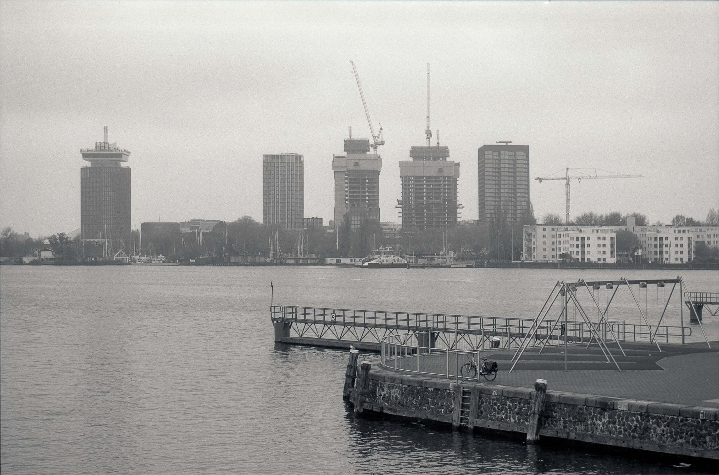 Uitzicht op een rivier met een stadsgezicht op de achtergrond, waaronder hoogbouw en bouwwerkzaamheden met hijskranen, en een weiland met een fiets en een schommel in de voorgrond.