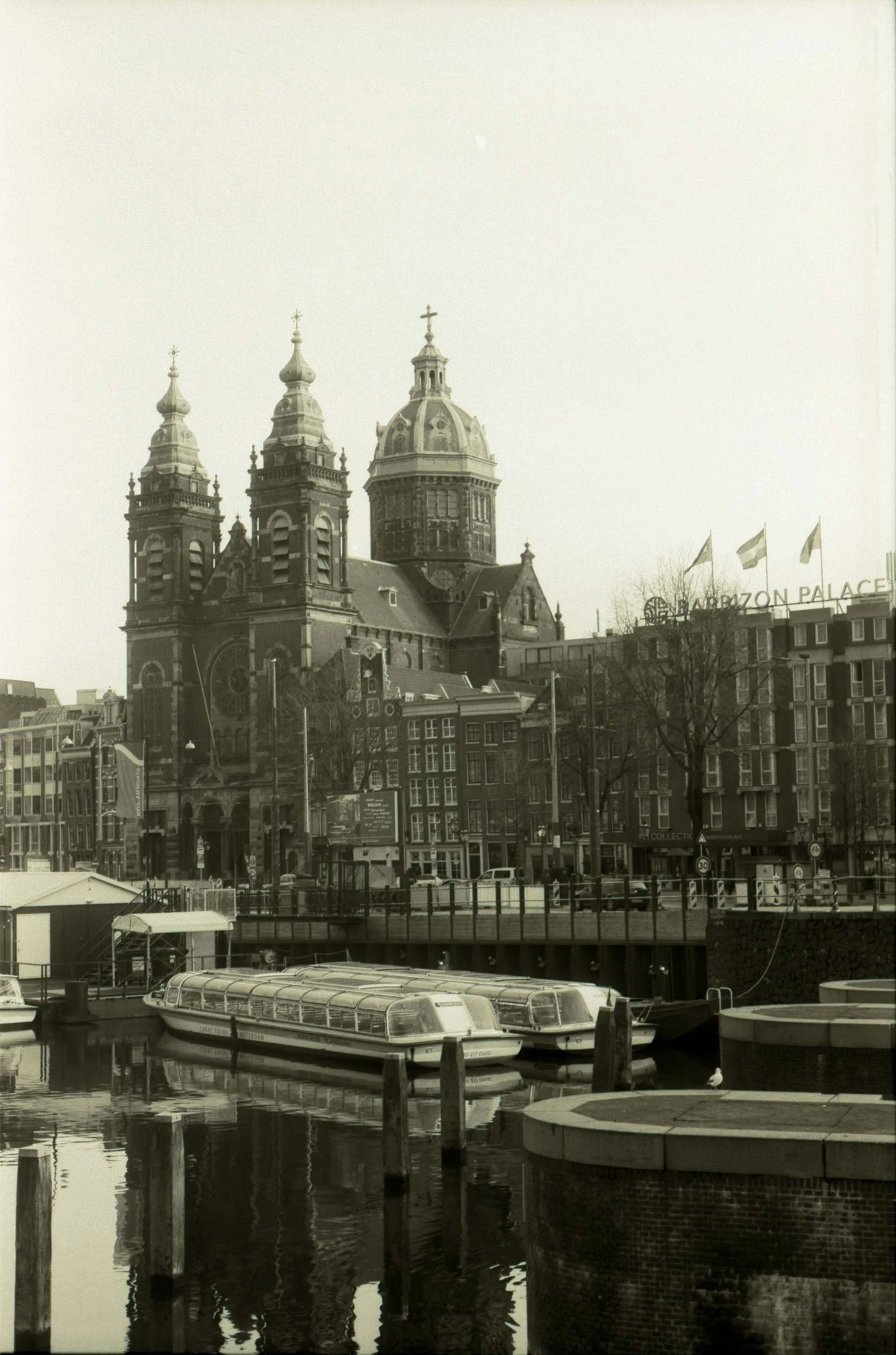Een zwart-wit foto van een grachtenstad met een kerk met drie torens op de achtergrond, vlakbij water met liggende boten en omringende gebouwen.