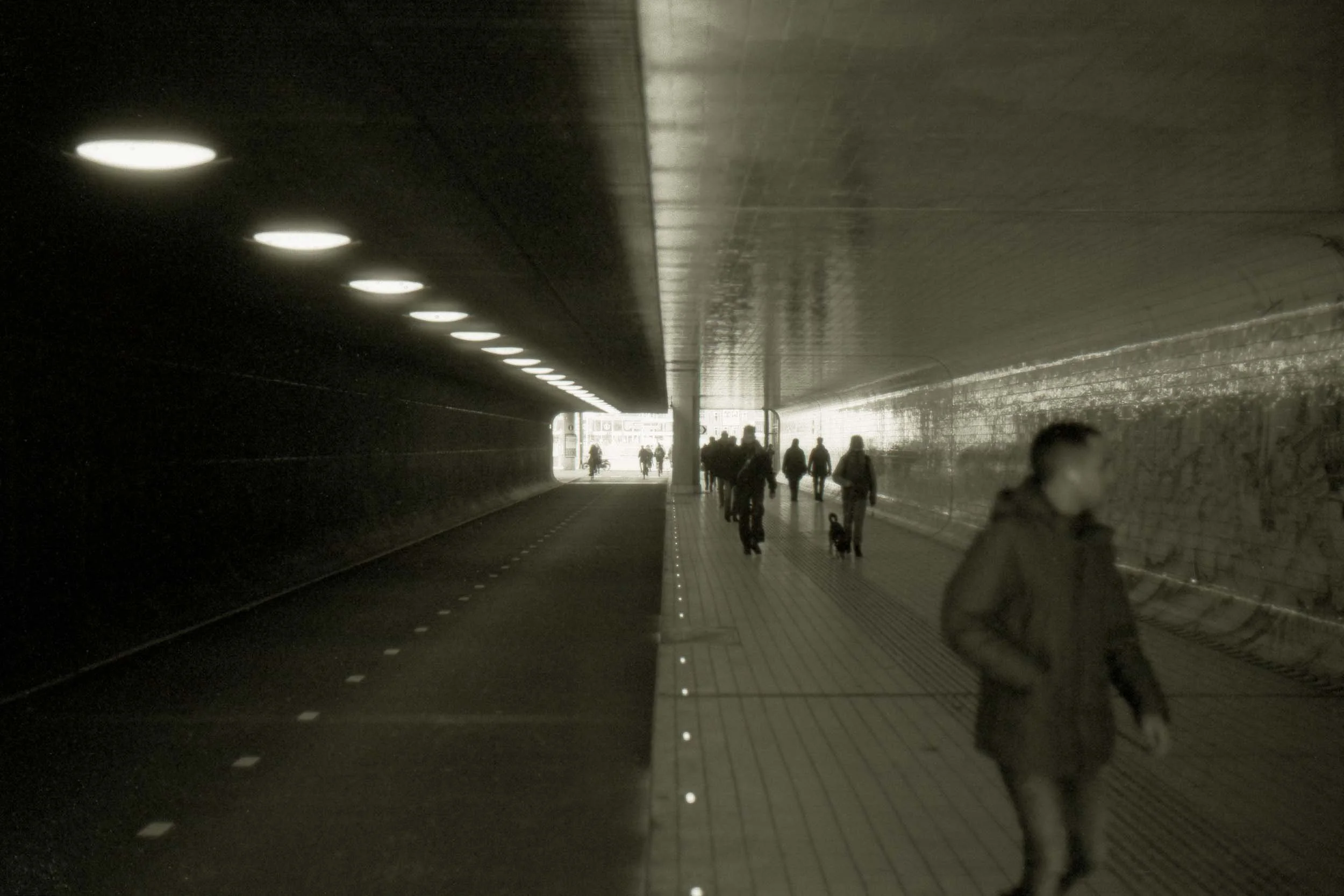 Mensen wandelen door een tunnel met verlichting aan het plafond, een deel van de wand is zwart en het andere deel is glanzend