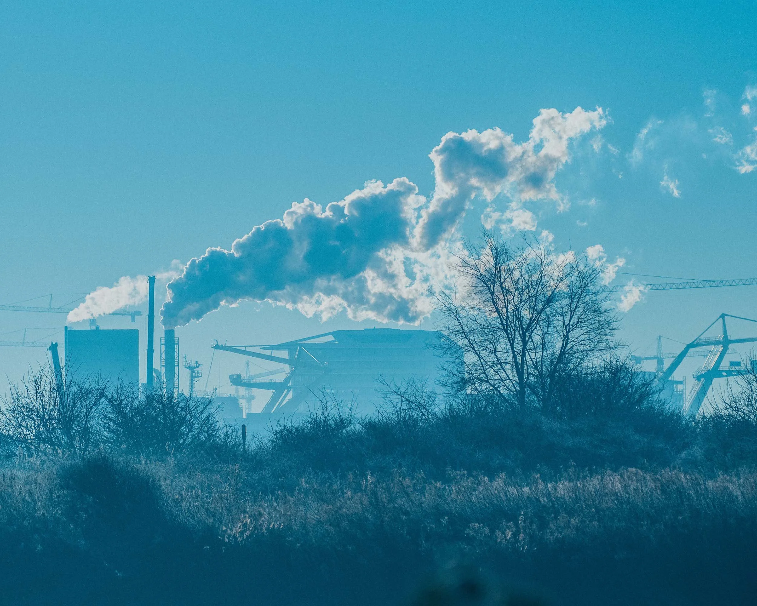 Een industriële fabriek die rook uit schoorstenen uitstoot, omgeven door bomen en bouwkranen onder een blauwe hemel.