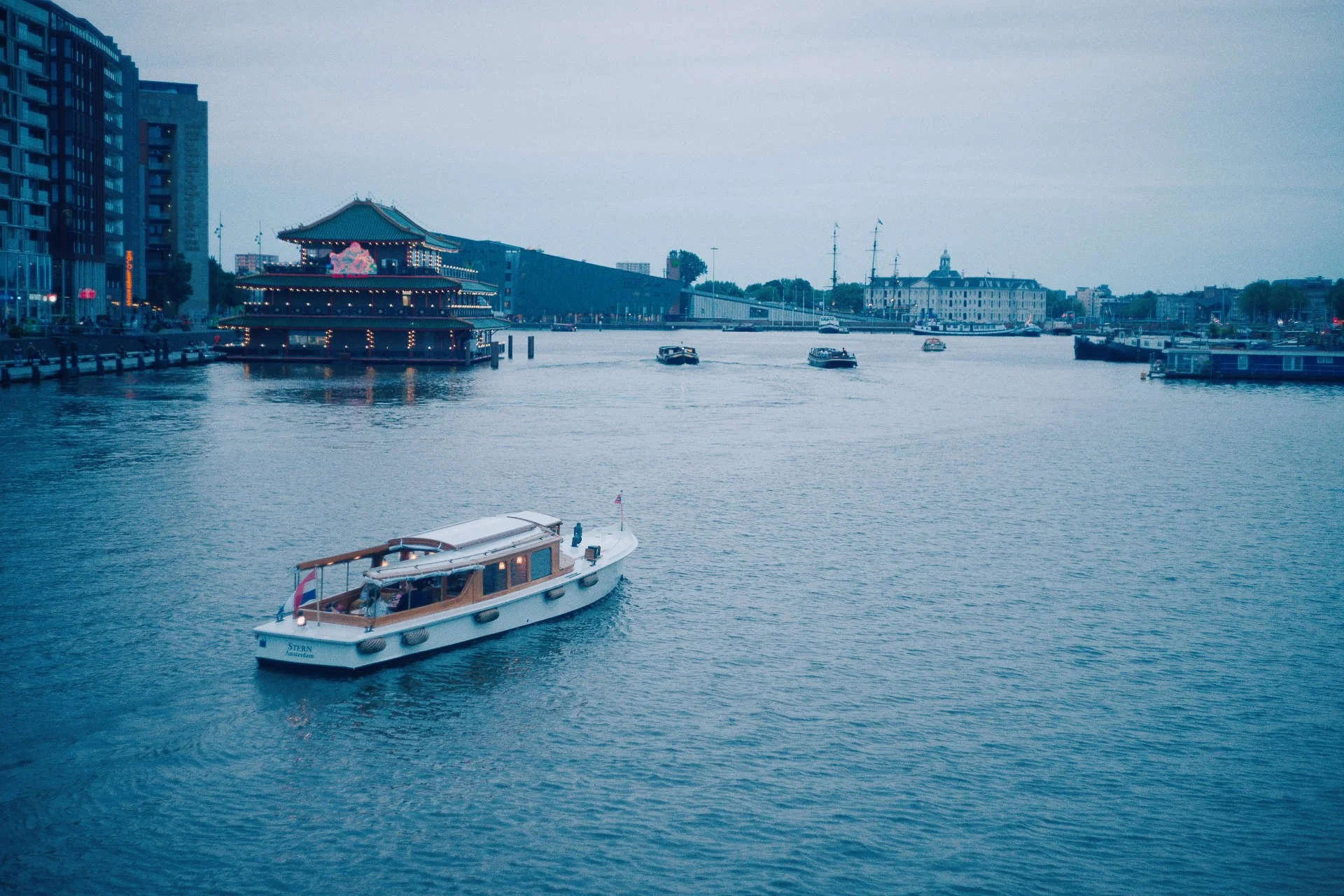 Een rivier met enkele boten, waaronder een Italiaanse rondvaartboot, omgeven door hoge moderne gebouwen en een traditioneel Chinees paviljoen met neonverlichting in een haven, tijdens de avond of schemer.