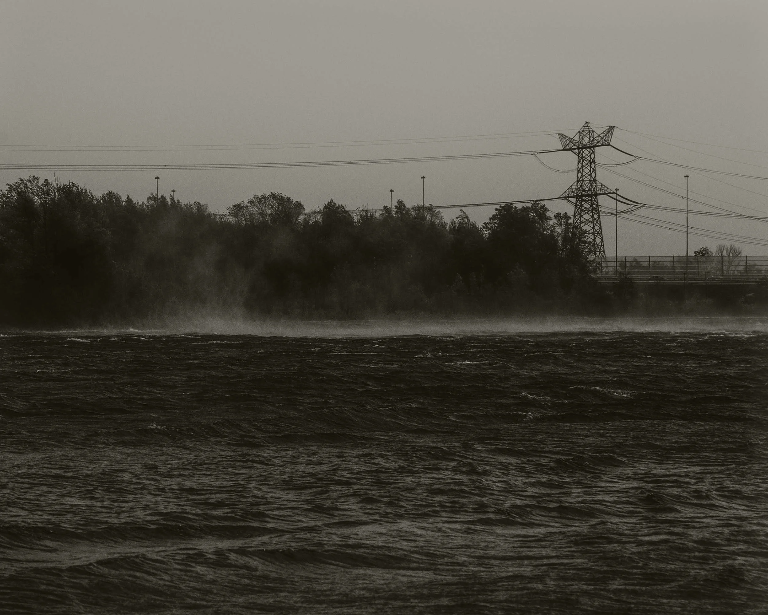 Een zwart-wit foto van een waterlichaam met rimpelingen, een bosrijke oever en een elektriciteitspaal met hoge spanningdraden in de verte.