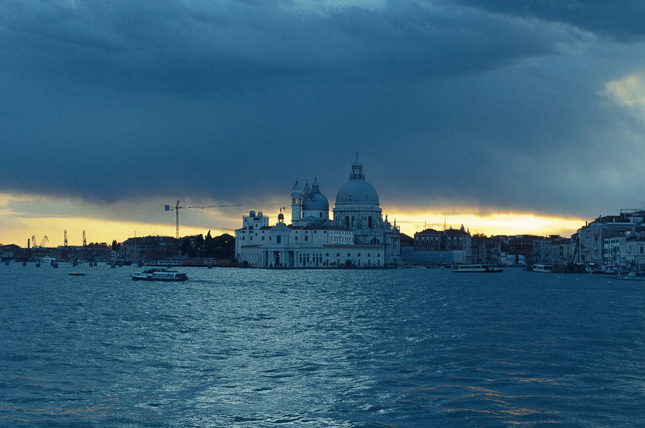 Uitzicht op de stad Venetië met de kerk Santa Maria della Salute op de voorgrond, onder een donkeren hemel bij zonsondergang, met boten op het water.