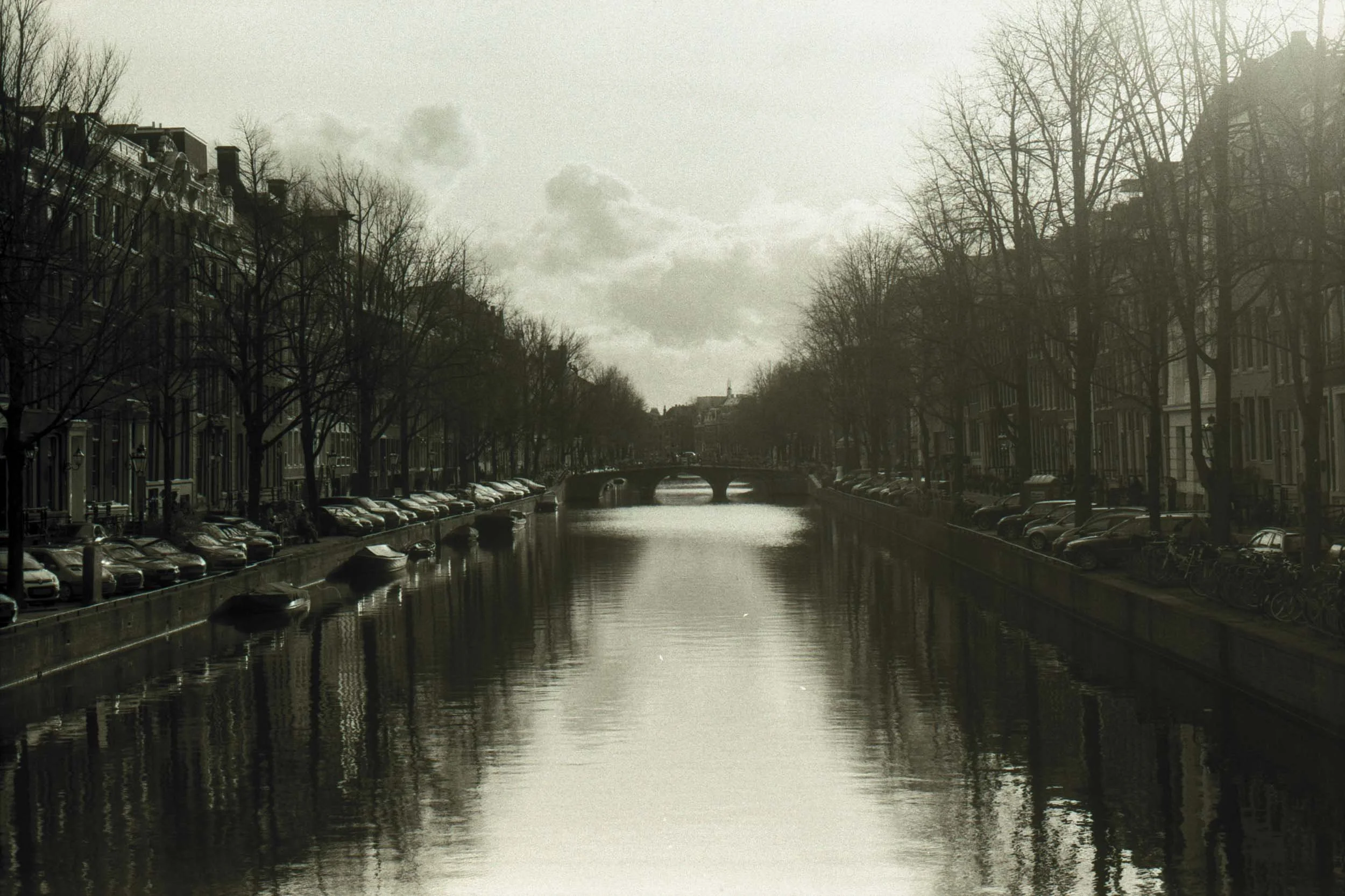 Een zwart-witafbeelding van een kanaal met oude gebouwen en bomen aan weerszijden, een brug in de verte en een bewolkte hemel.
