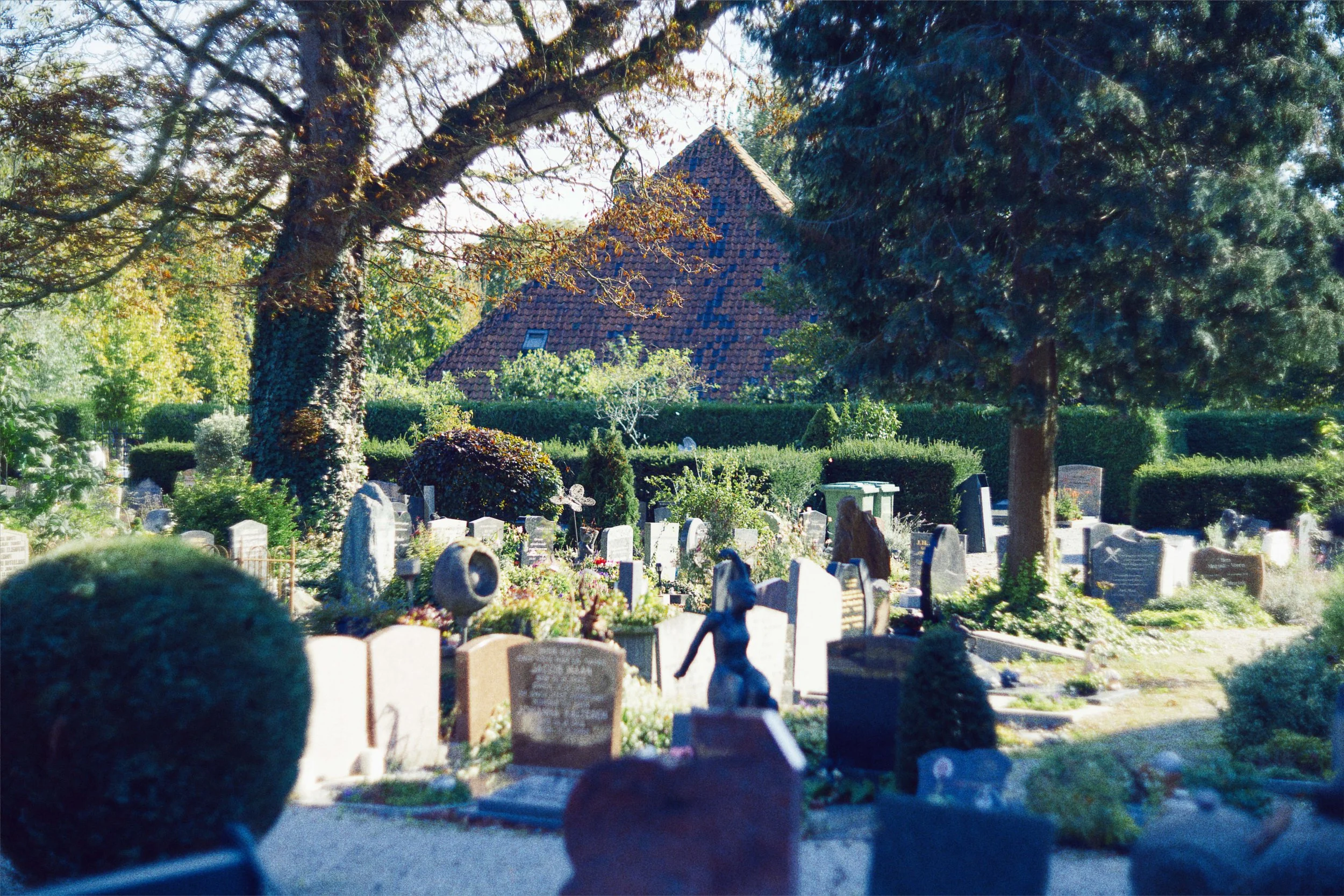 Een begraafplaats met graven, bomen en een huis op de achtergrond. Het lijkt een rustige en groene omgeving.