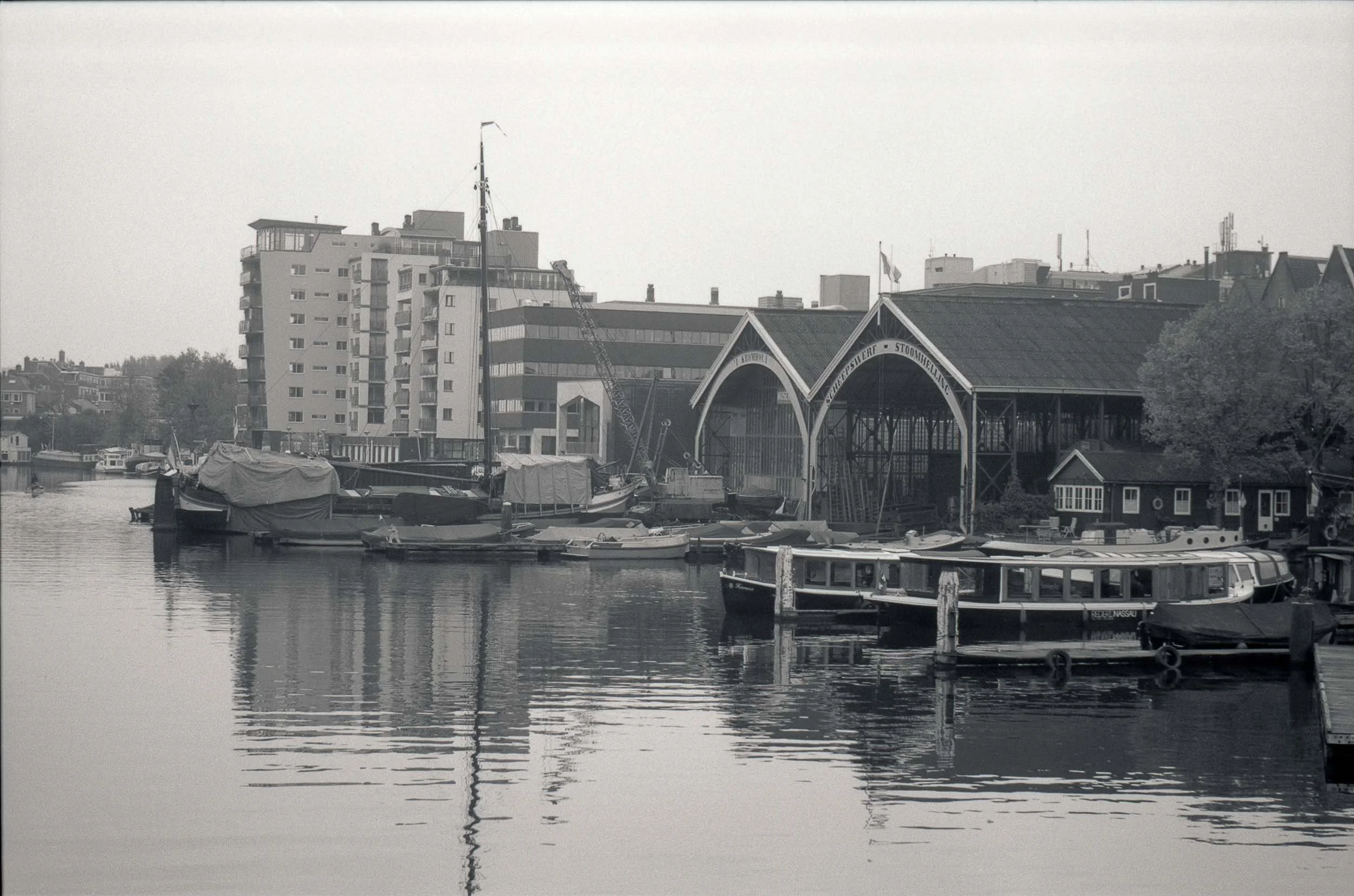 Een zwart-witfoto van een haven met verschillende boten en schepen, en gebouwen op de achtergrond.