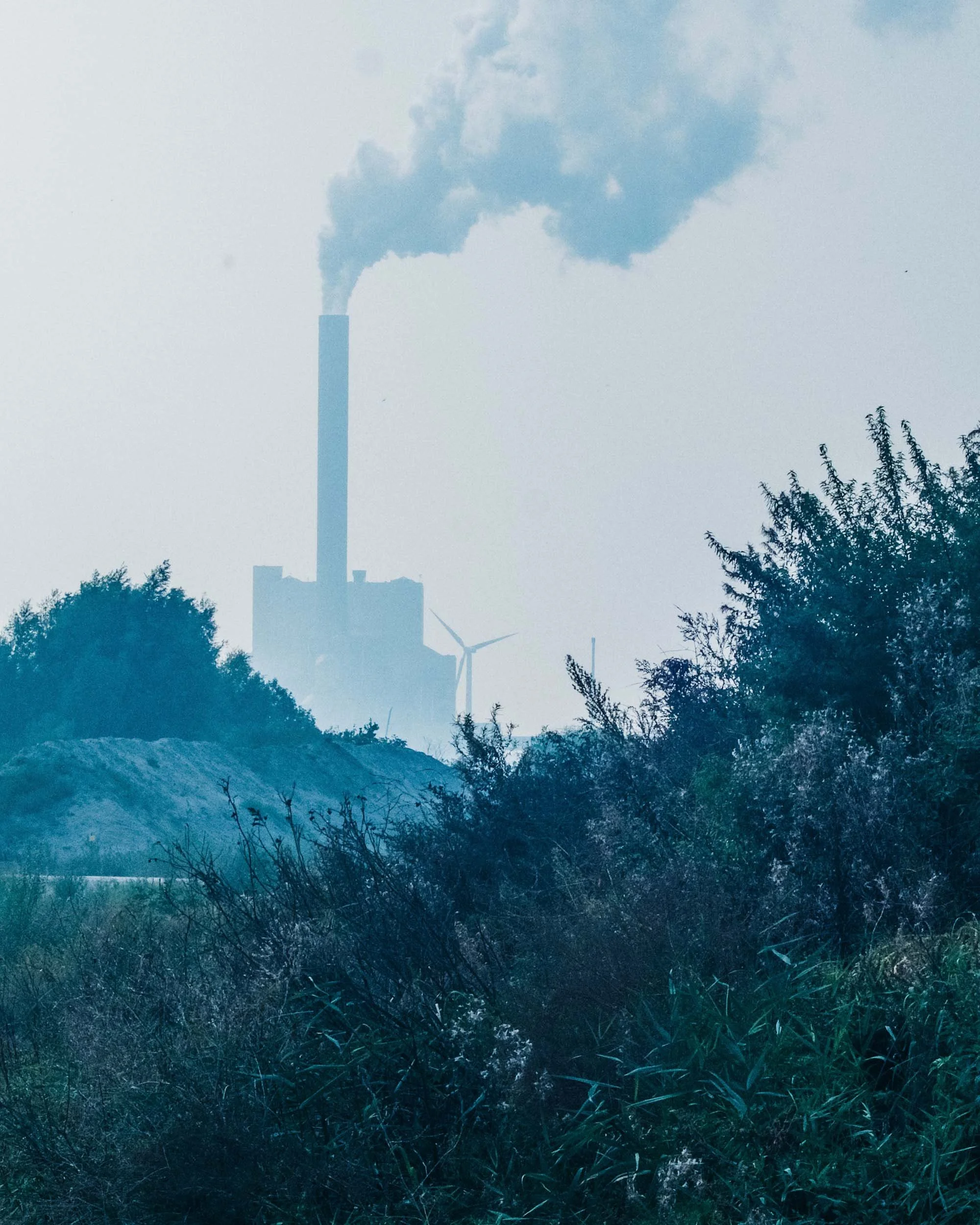 Een industriële fabriek met een schoorsteen die roet uitstoot, omringd door bomen en windturbines.