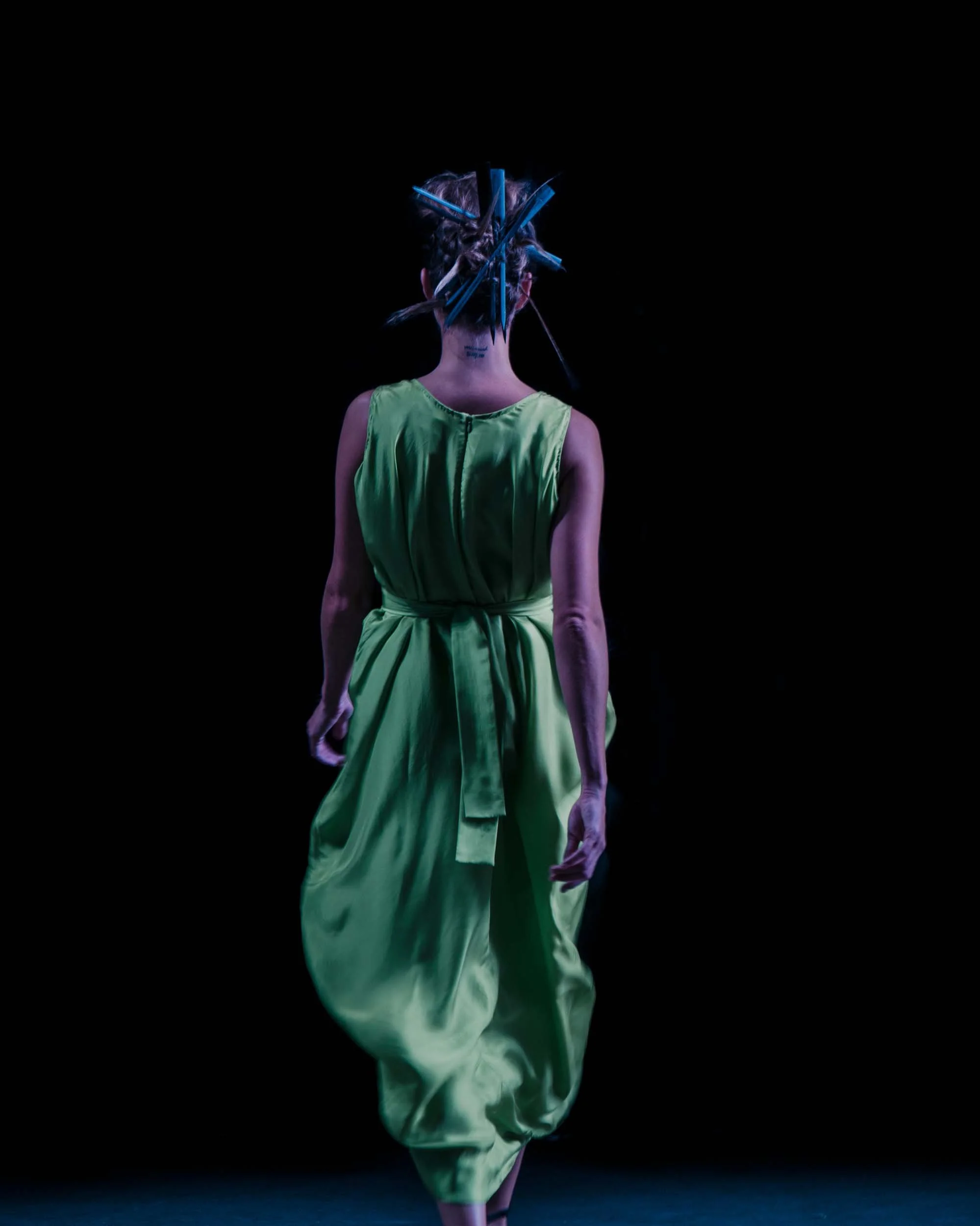 Een vrouw met een groene jurk en een kunstzinnig kapsel dat bestaat uit vele blauwe stiften, die in een donkere ruimte loopt.