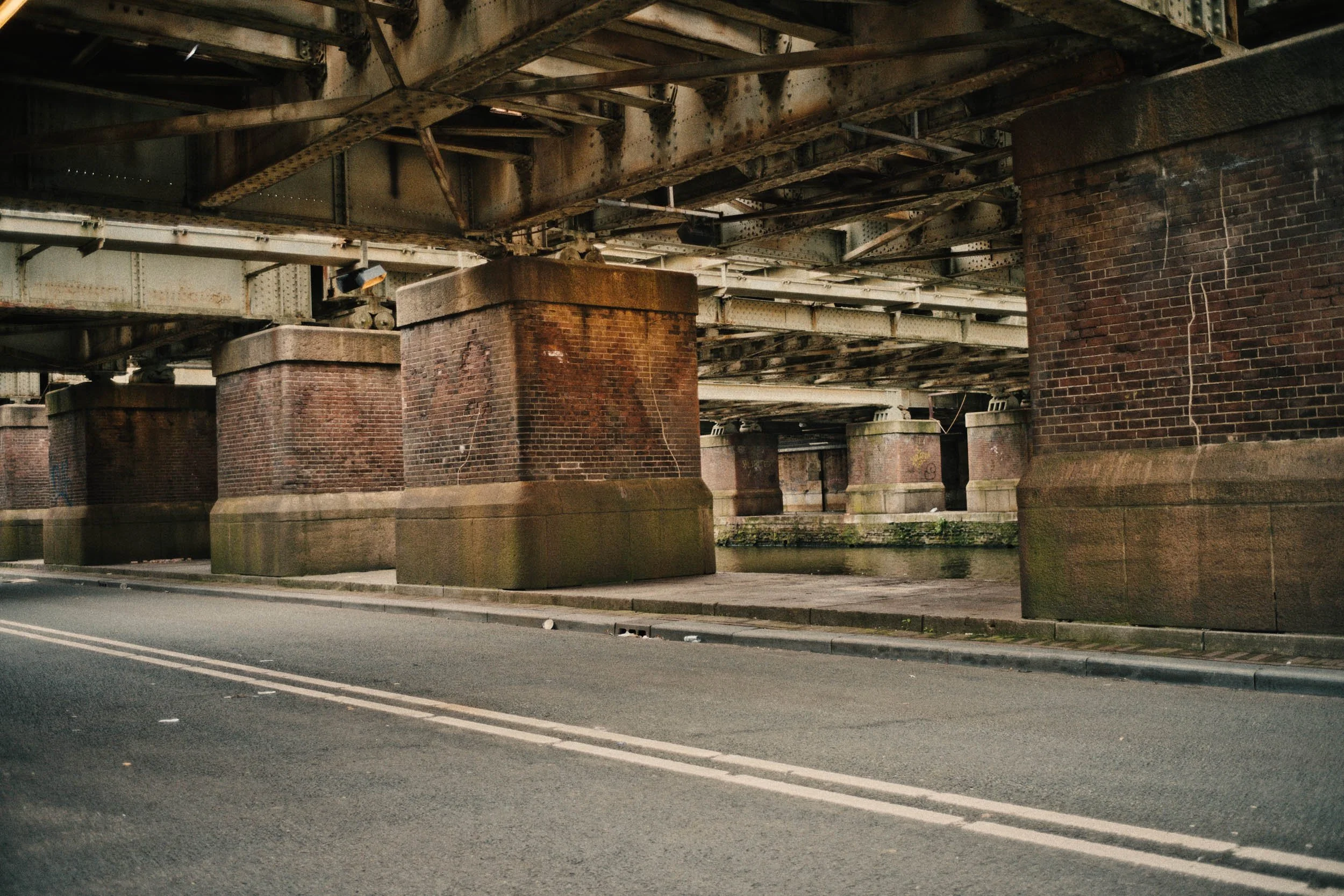 Onder een spoorbrug zien we een straat met een dubbele witte lijn en stenen brugpijlers van rood baksteen en beton onder de brug, met een waterloop eronder.