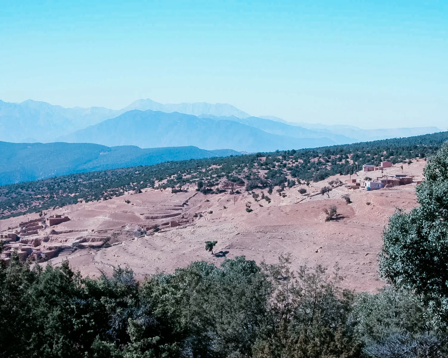 Een berglandschap met enkele kleine dorpen, bomen en een enkele schaap in de voorgrond, en bergen in de verte onder een heldere blauwe hemel.