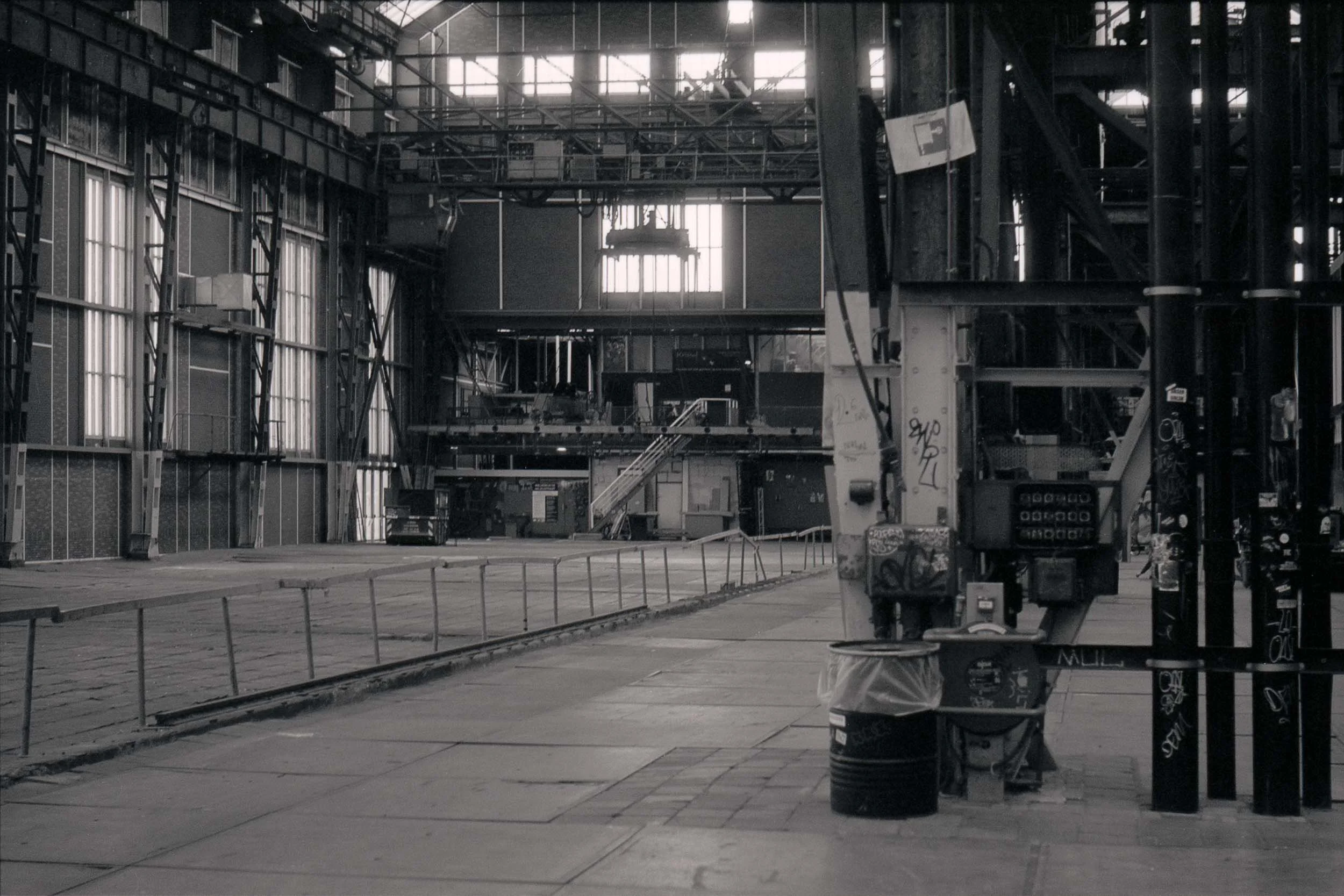 Leeg industrieel gebouw met grote ramen en metalen structuur, geen mensen, sommige graffiti op muren.