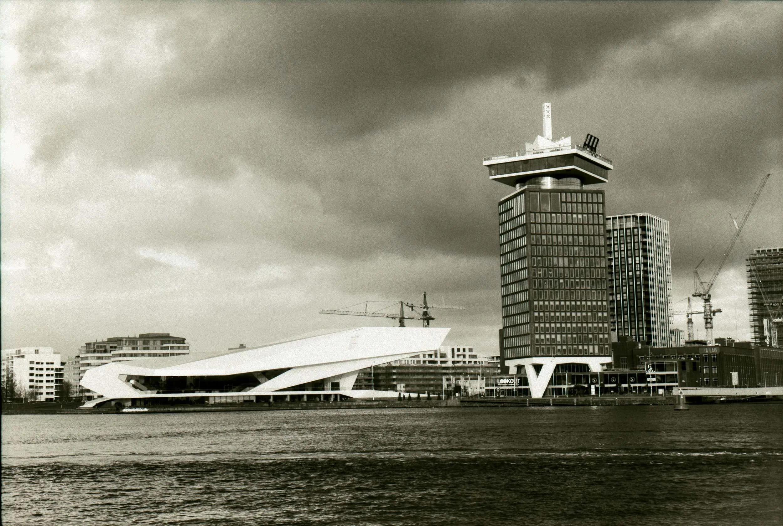 Zwart-wit foto van een moderne skyline met een opvallende hoge toren en een futuristisch gebouw, water in de voorgrond en een donker dreigend wolkendek.
