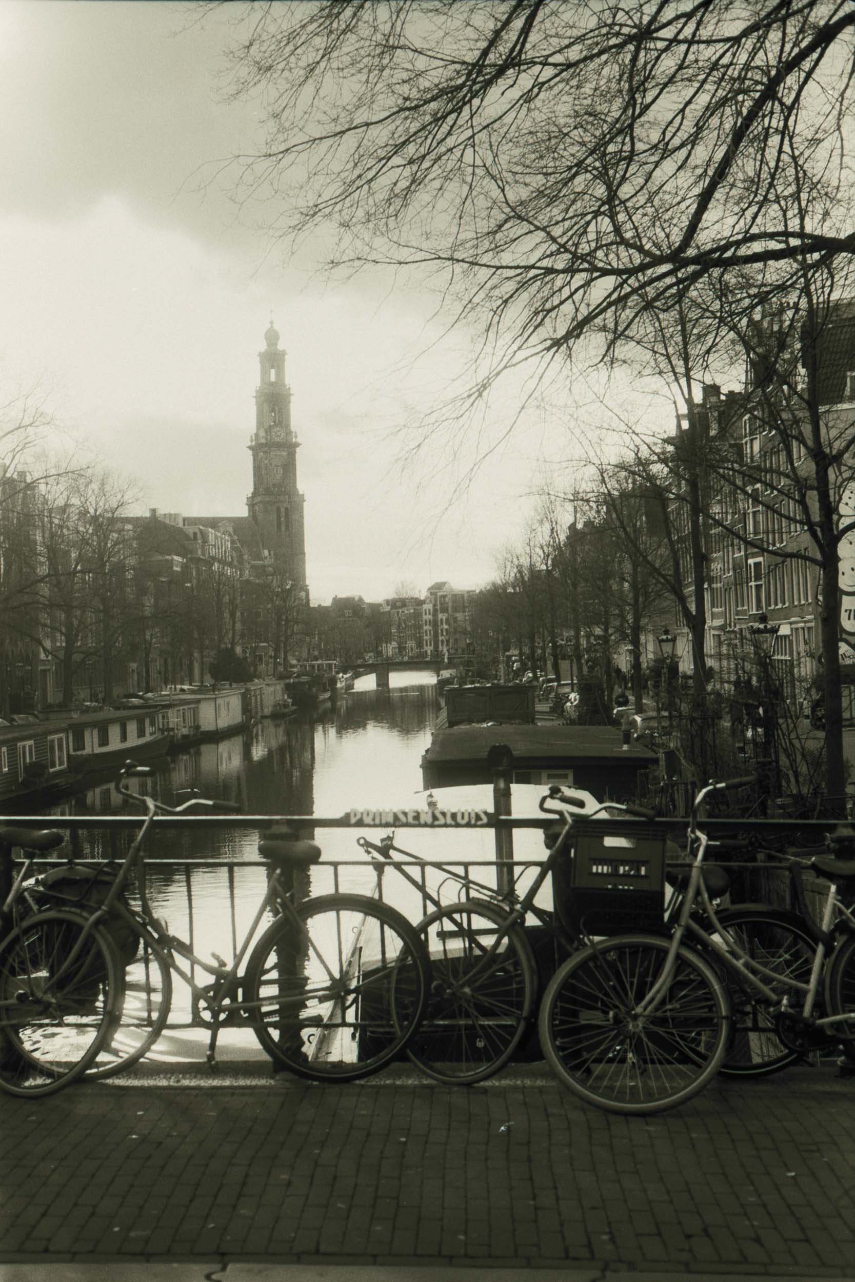 Beboste fietsen aan een brug over een kanaal in een stad, met historische gebouwen en een toren, in zwart-wit.
