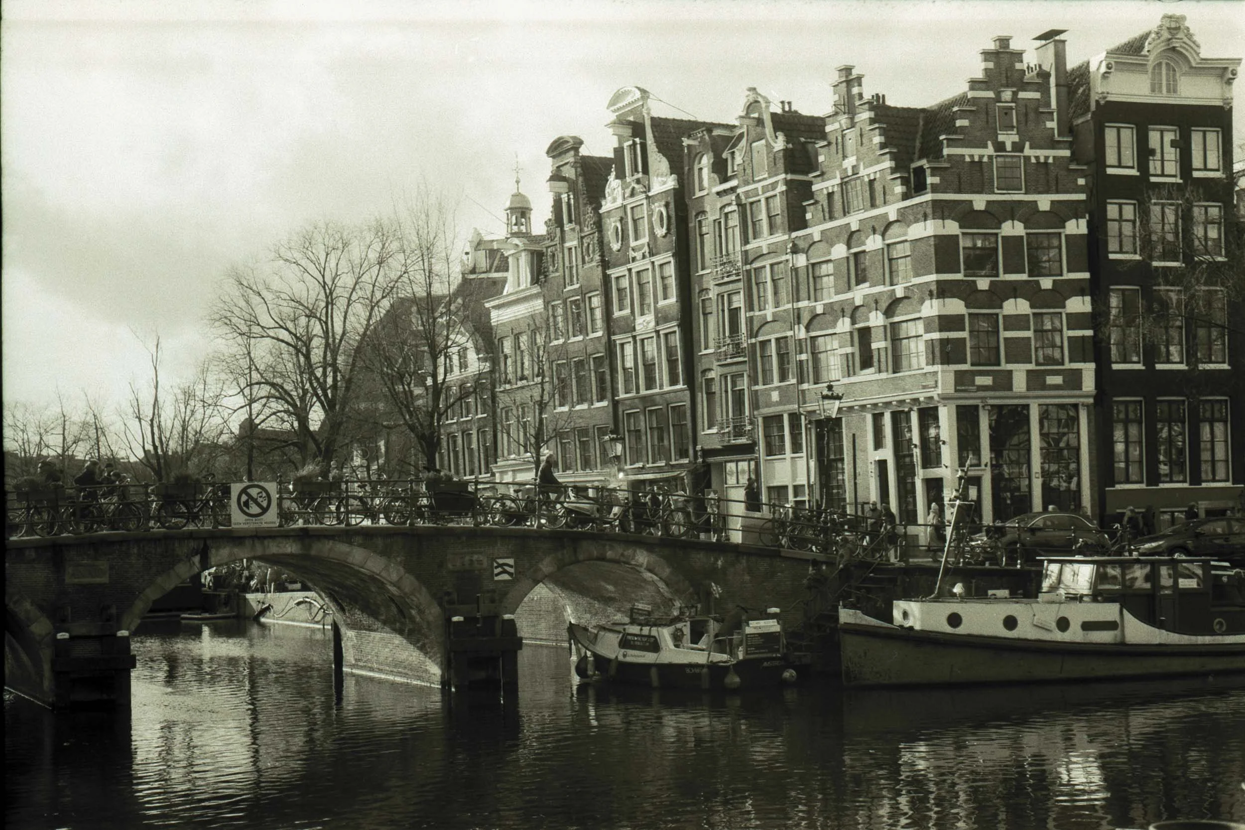 Een zwart-wit foto van een grachtenpand in Nederland met een brug en boten in de grachten, bomen zonder bladeren en een drukke straat met fietsen en auto's.