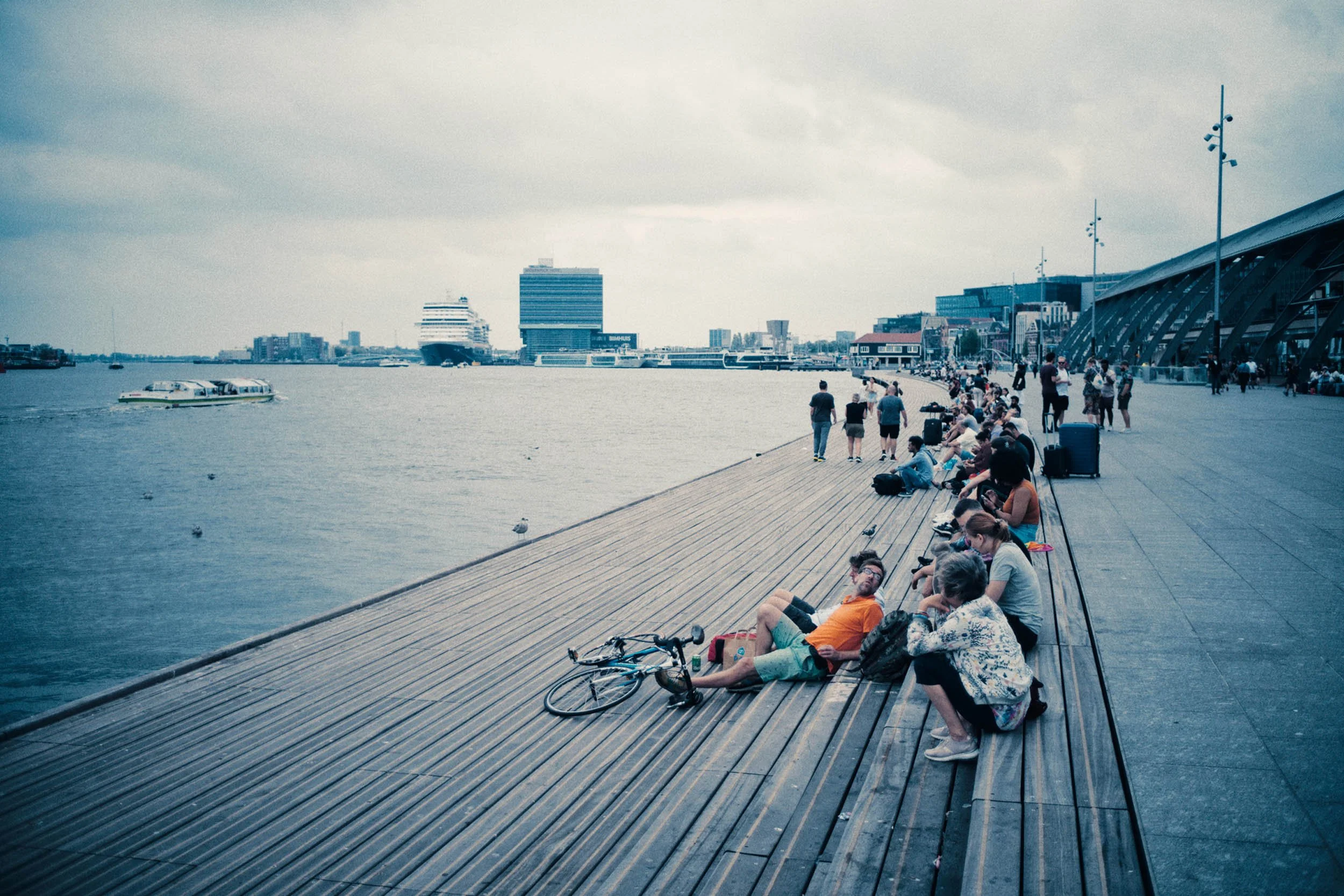 Mensen zitten en wandelen langs een promenade aan het water, met boten en moderne gebouwen op de achtergrond.